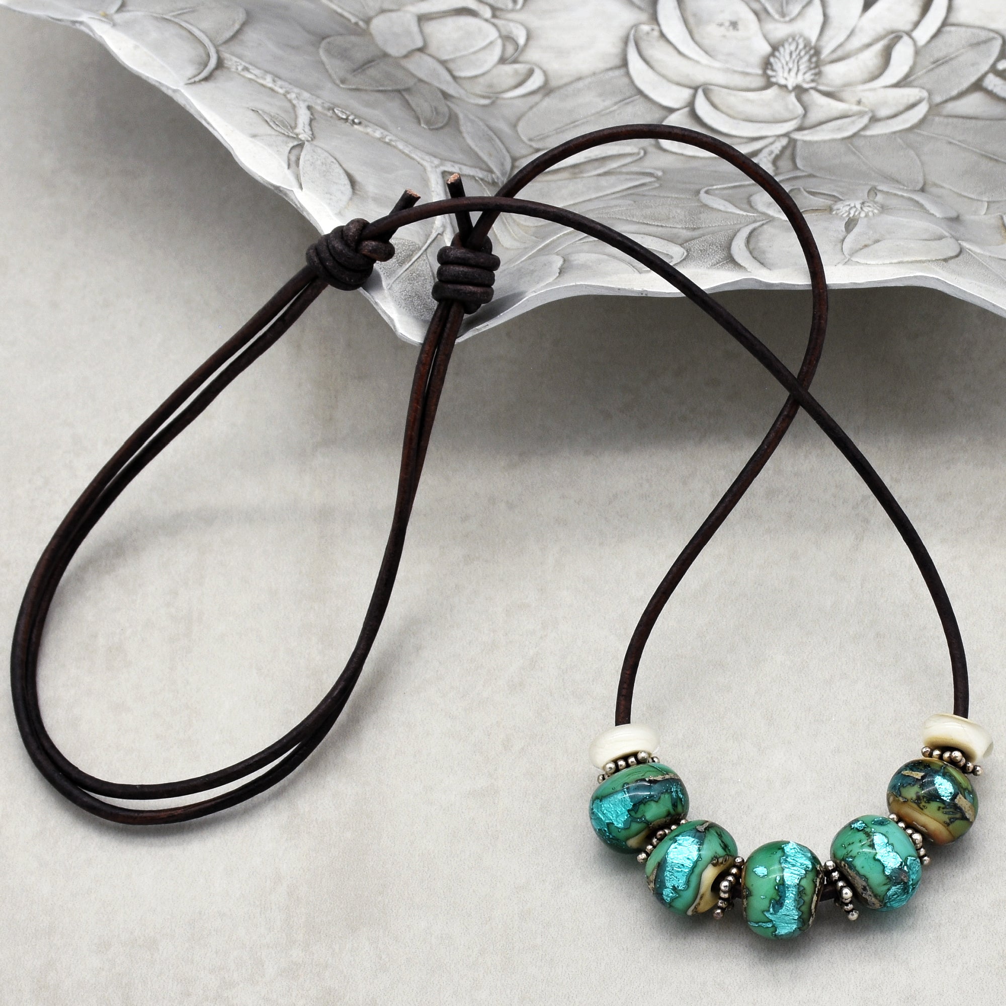 This image shows a necklace. It consists of a dark brown cord with several turquoise and metallic beads strung on it. The knots at the ends suggest it is designed to be worn around the neck.