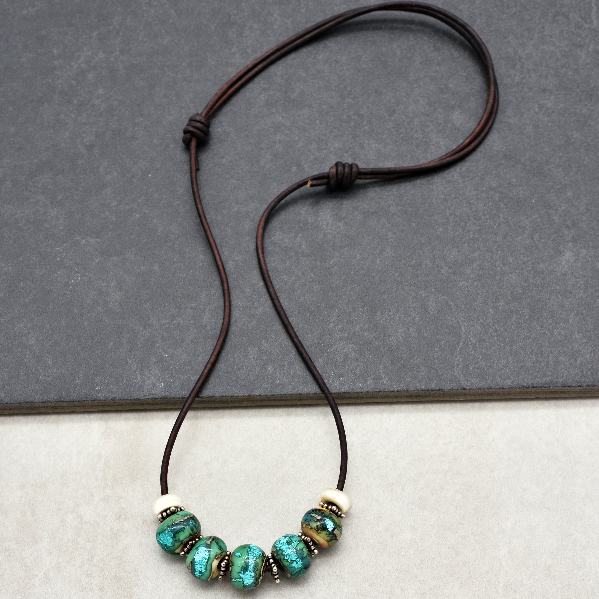 This image shows a necklace. It consists of a brown cord with five beads strung onto it. The beads are predominantly turquoise with some metallic and white accent beads in between. The style, length, and arrangement are consistent with a necklace rather than an earring or bracelet.