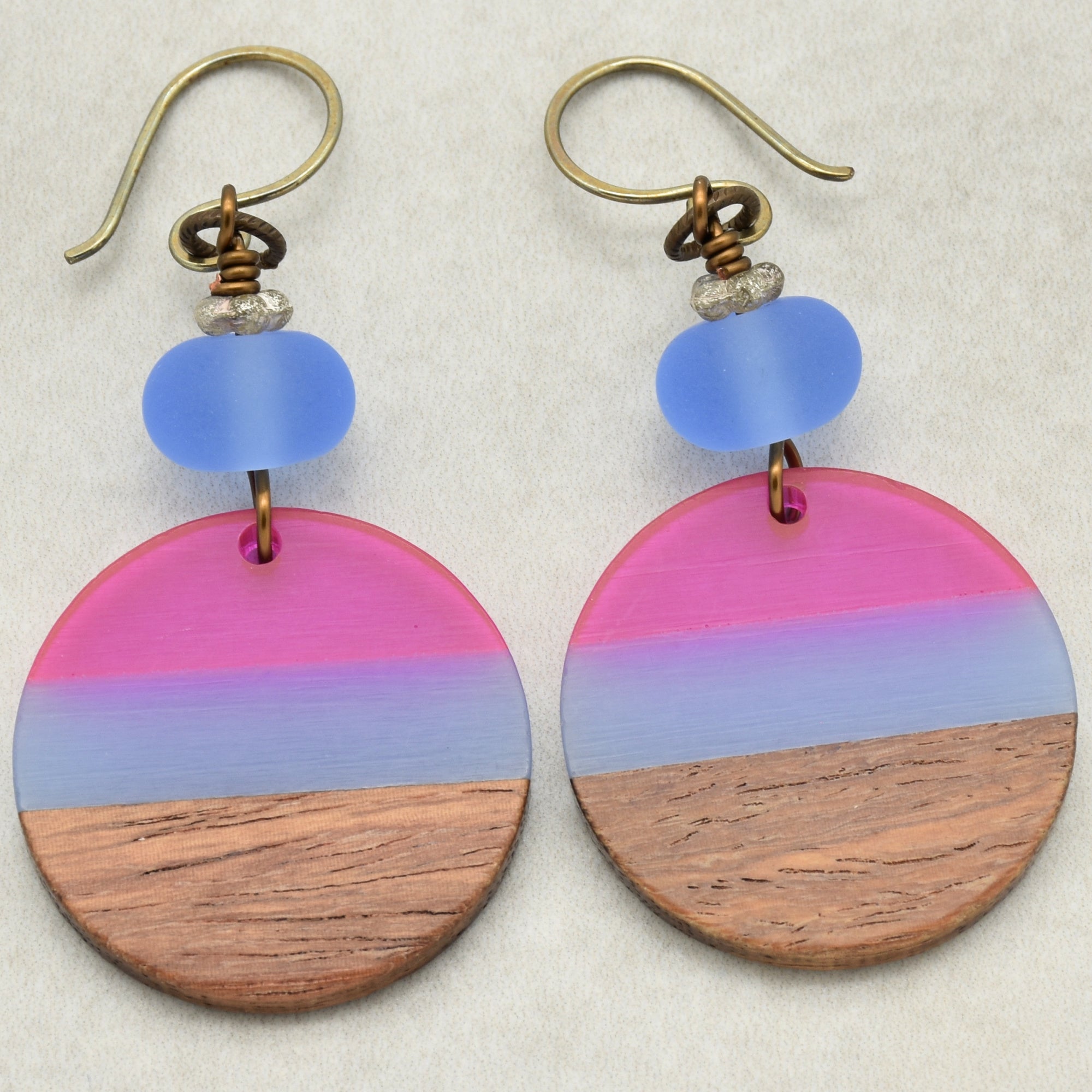 The image shows a pair of earrings. They have metal hooks and feature round pendants with a design that includes blue and pink colors above a section that looks like wood. There is also a small blue bead above each round pendant.