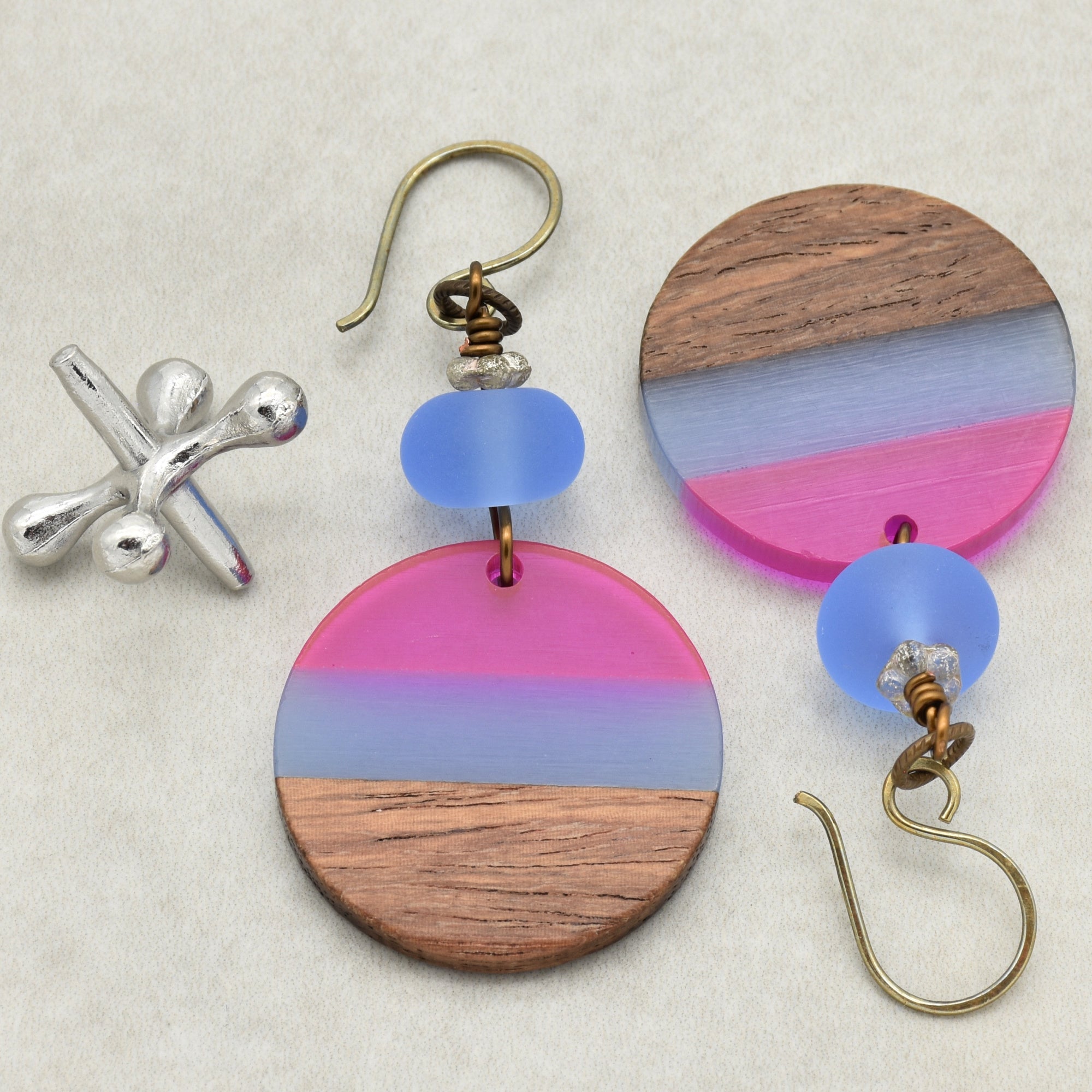 The image shows a pair of earrings. They have a hook for the ear and a round decorative element featuring wood and colored sections in pink and blue, with a blue bead above the round piece.