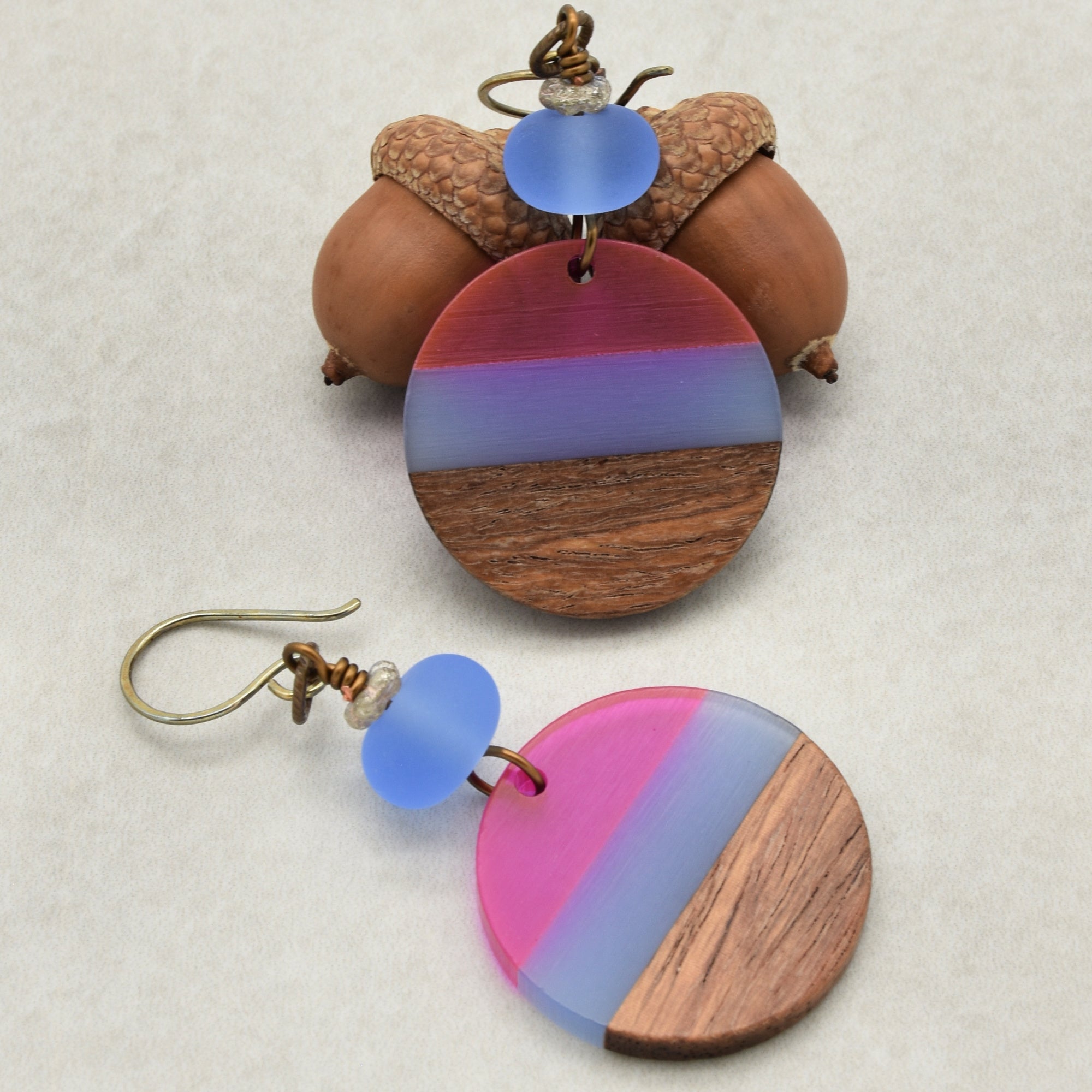 The image shows a pair of earrings. They are circular with a combination of wood and colored materials (pink, purple, and blue). Each earring has a hook for wearing in pierced ears.