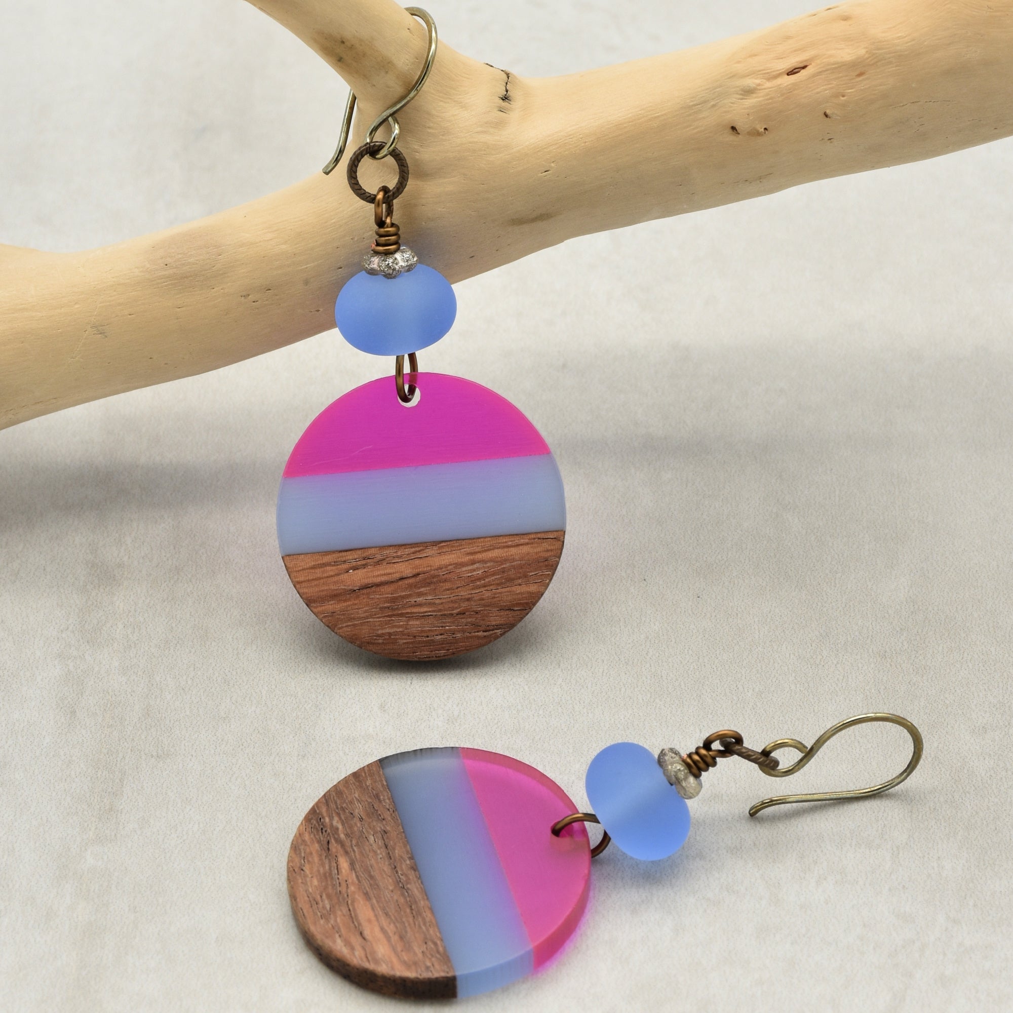 The image shows a pair of earrings. They have a circular design with sections of pink, translucent white, and wood, along with a round blue bead above the circular piece. The earrings have hooks for wearing.