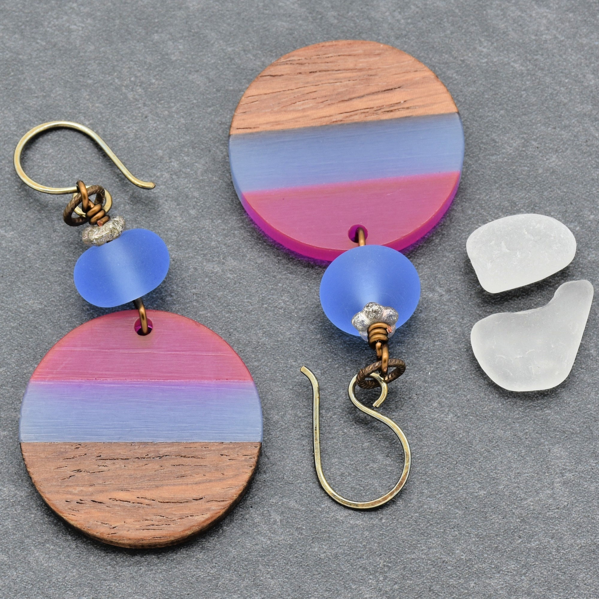 This image shows a pair of earrings. The jewelry pieces have hooks typical of earrings, and each features a round pendant with a gradient of pink, blue, and wood tones, adorned with a blue bead and a small textured silver accent above the pendant.