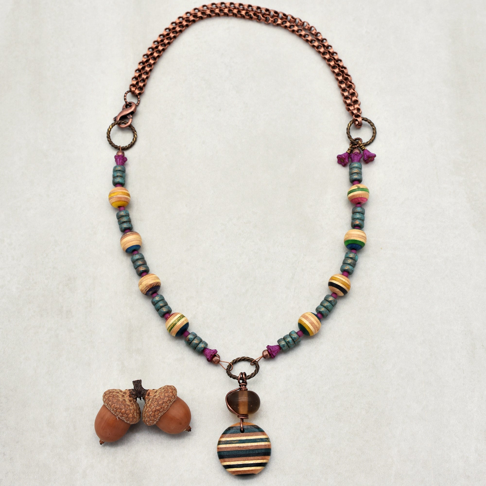 This image shows a necklace. The necklace features a copper chain, colorful striped beads, and a round pendant with a striped pattern. There are also some purple and blue beads along its length, and it is displayed alongside two acorns.