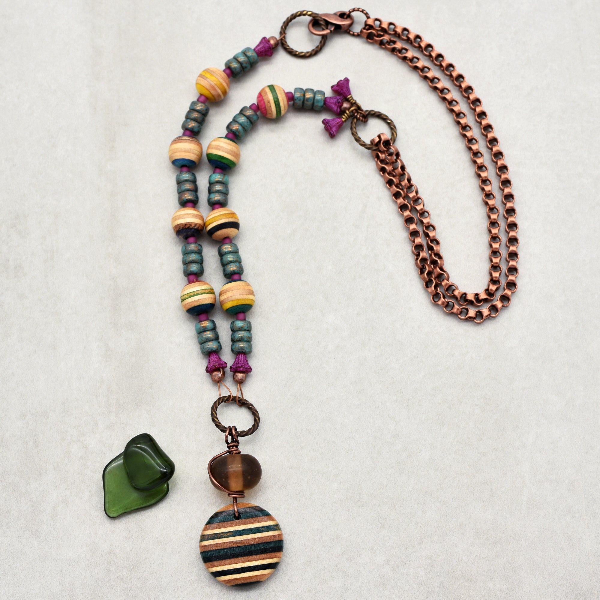 This image shows a necklace. It consists of a copper-colored chain and a section with various beads in colors like blue, yellow, purple, and natural wood tones. It also has a striped circular pendant and an additional green glass piece. The length and design indicate it is a necklace rather than an earring or bracelet.