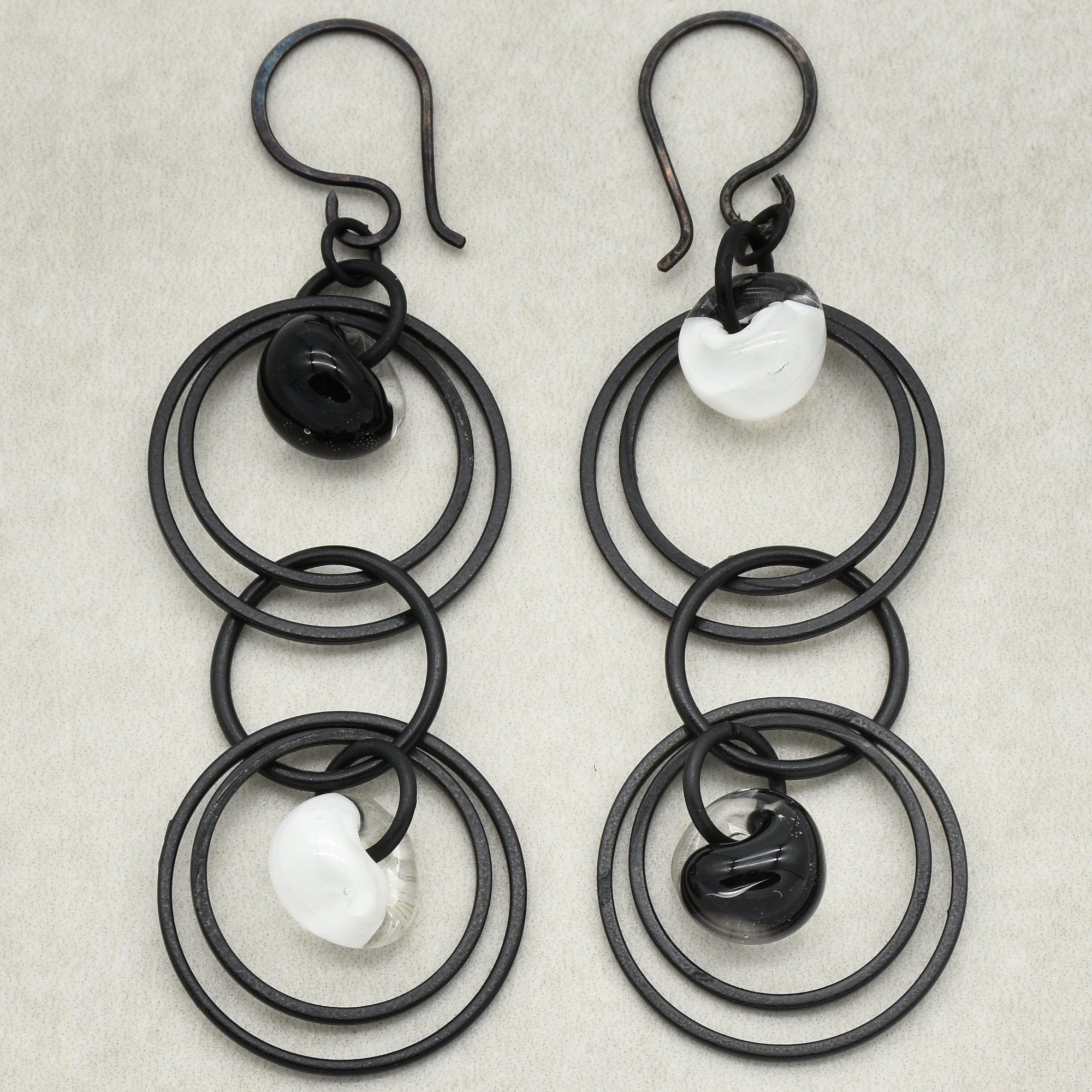 This image shows a pair of earrings. The jewelry has hooks at the top, which are characteristic of earrings designed to be worn on pierced ears. The design features multiple interlinked black rings with black and white glass or bead accents.