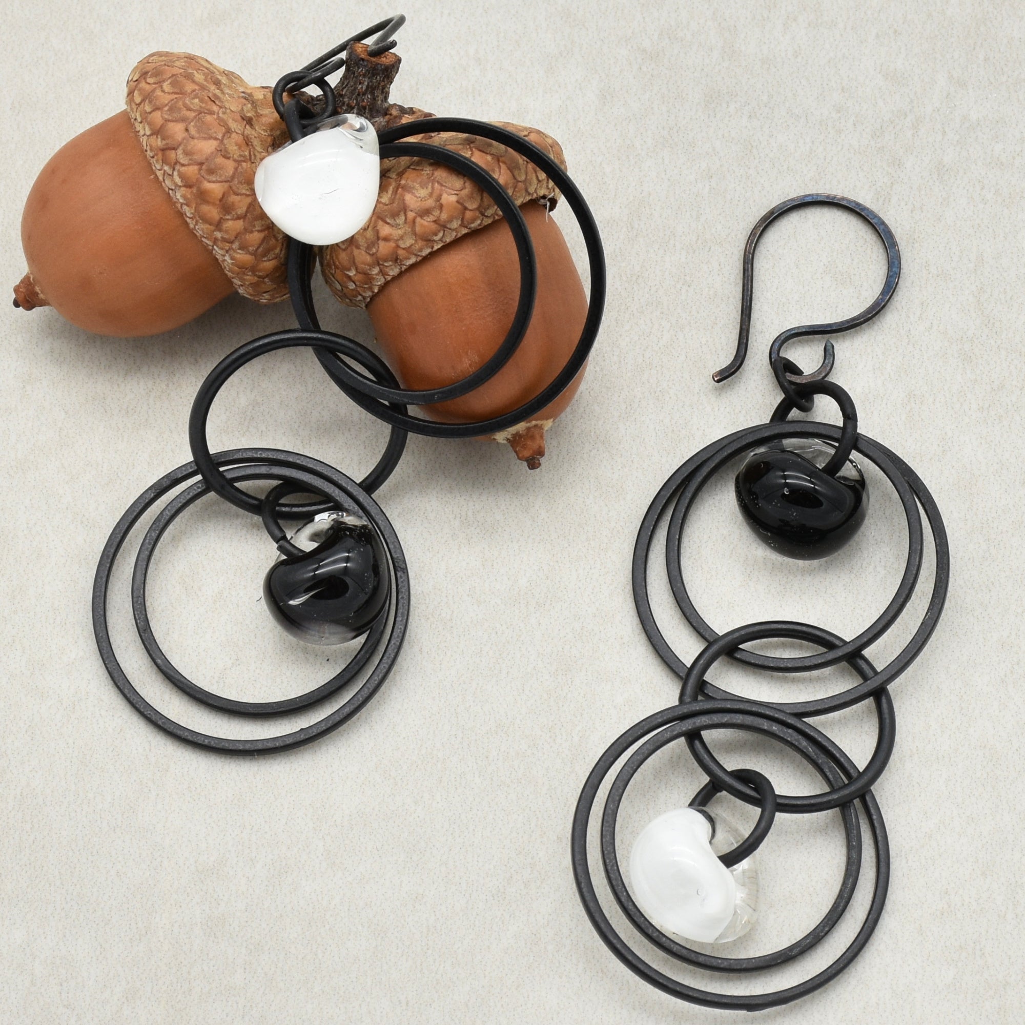 The image shows a pair of earrings. They have multiple black circular hoops linked together with small glass beads, one white and one black, attached within the hoops. One earring is hanging on a pair of acorns.