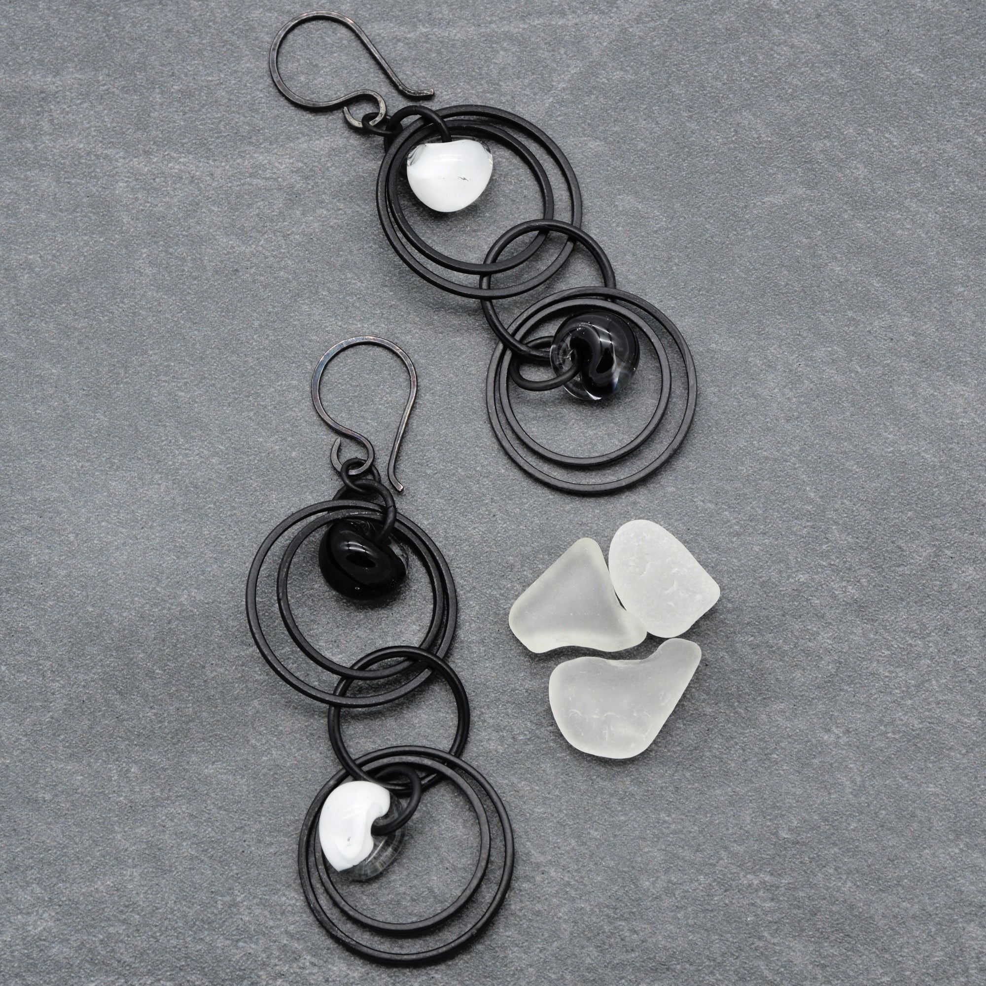 The image shows a pair of earrings. They have black circular metal components with white and black sea glass or stone accents. The hooks at the top of the items indicate that they are earrings.