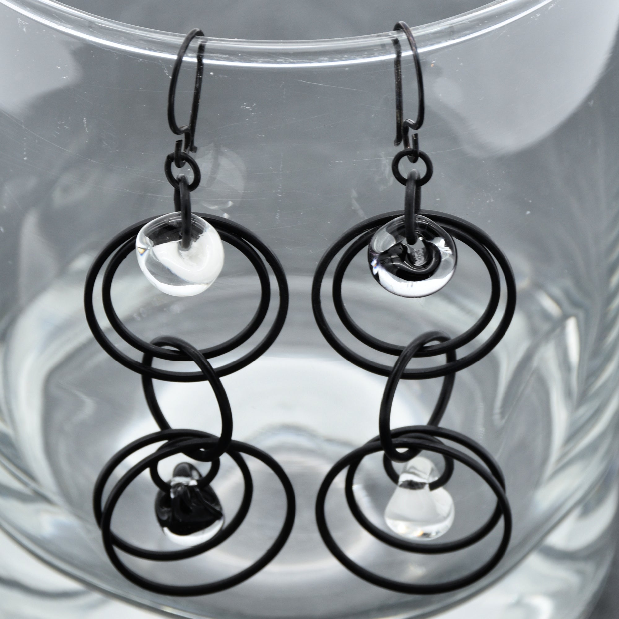 The image shows a pair of earrings. They have multiple black circular loops with small clear glass or crystal beads hanging in the center of some loops. The earrings are hanging on a transparent surface.