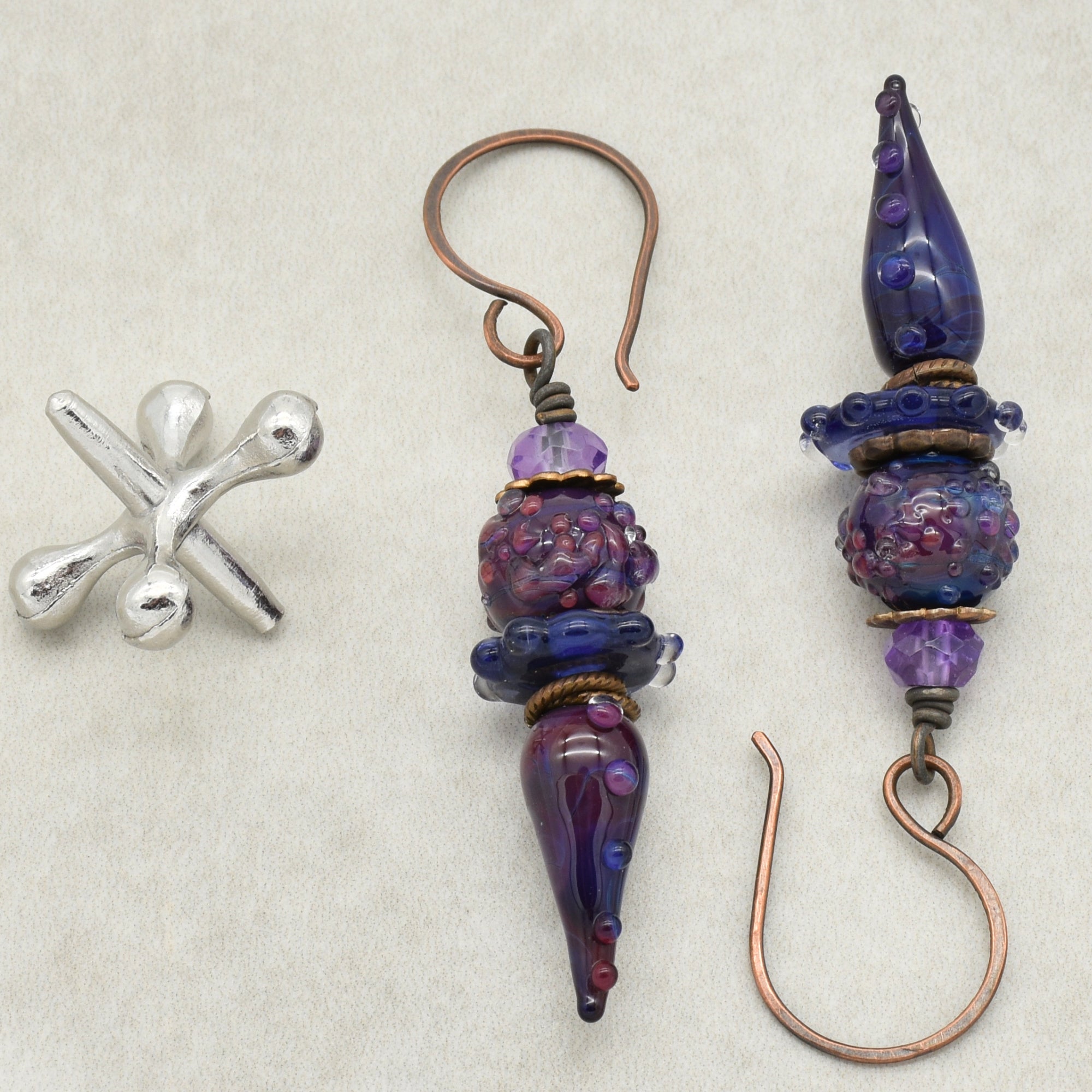 The image shows a pair of earrings. The jewelry pieces have hooks attached, which are typically characteristic of earrings. The design includes purple and blue beads with decorative elements and copper-colored hooks. There is also a small silver-colored decorative piece on the left, but the primary jewelry items with hooks are earrings.