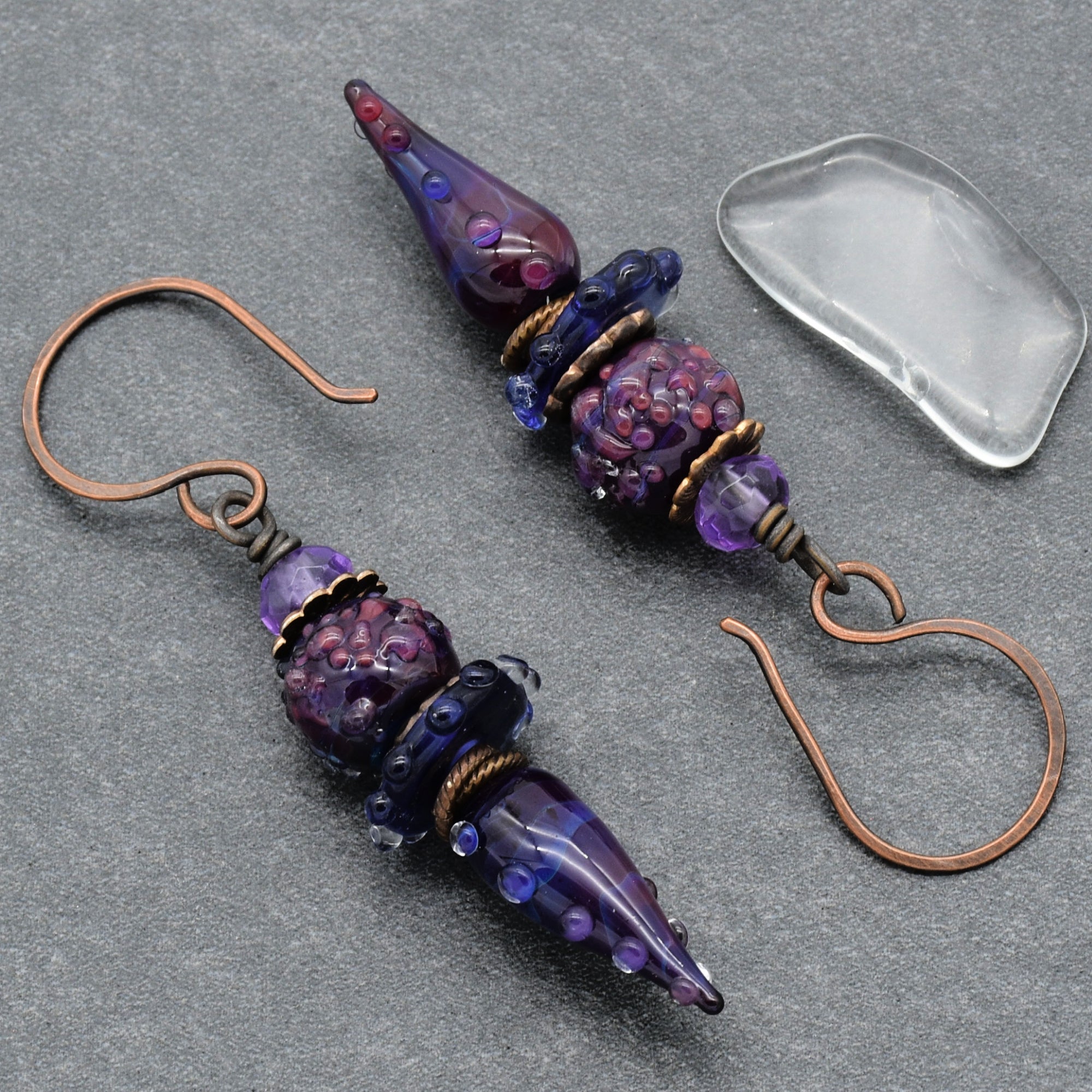 The image shows a pair of earrings. They have hooks for inserting into pierced ears and are adorned with decorative beads in shades of purple and blue.