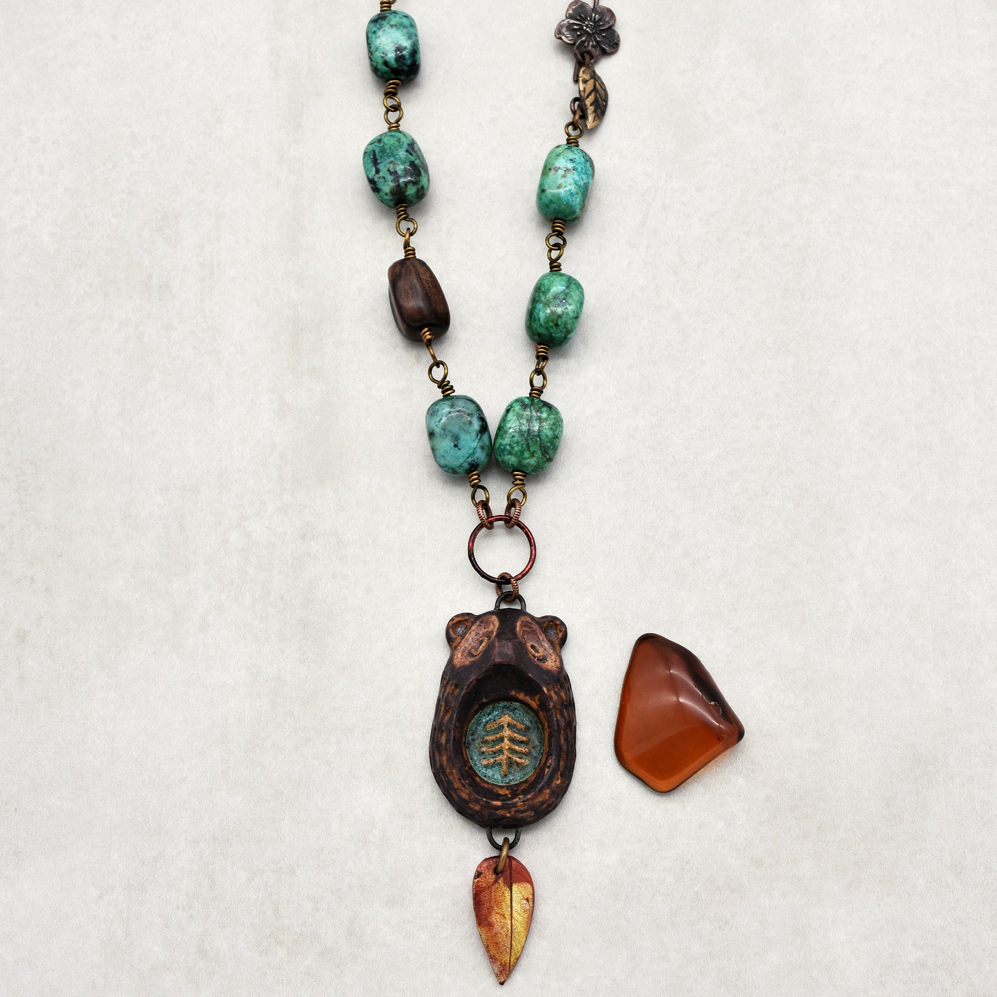 This image shows a necklace. The necklace features turquoise and wooden beads, with a pendant that has a bear design and a leaf ornament hanging from it. There is also an additional polished stone to the side.
