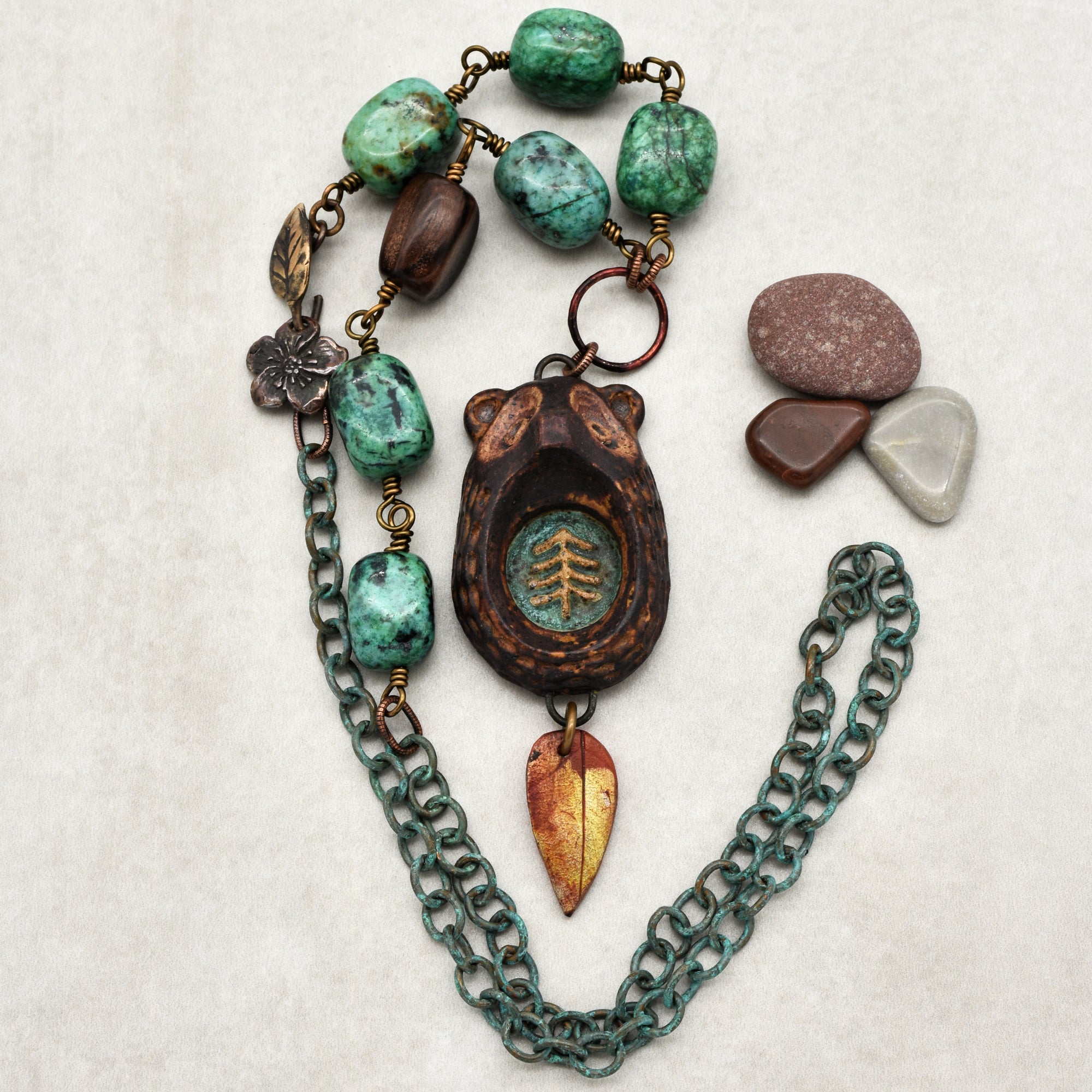This image shows a necklace. It features large turquoise stones, a bear-shaped pendant with an etched tree symbol, and a leaf-shaped charm. The necklace also has a chain with a greenish patina and smaller decorative elements like a flower and leaf charm.