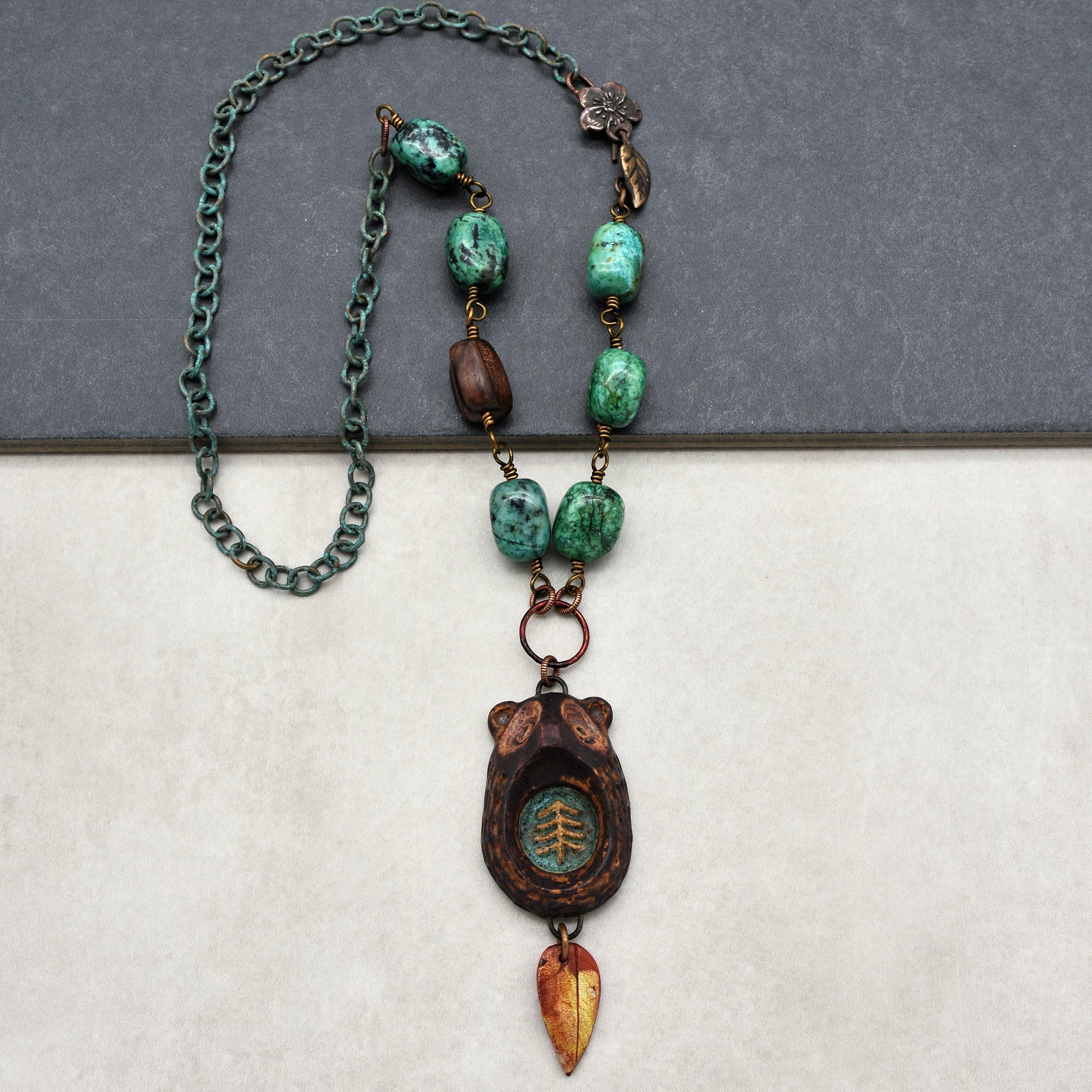 The image shows a necklace. It features a chain with green and brown beads, and a pendant shaped like a bear with a tree design inside, along with a small leaf charm hanging below it.