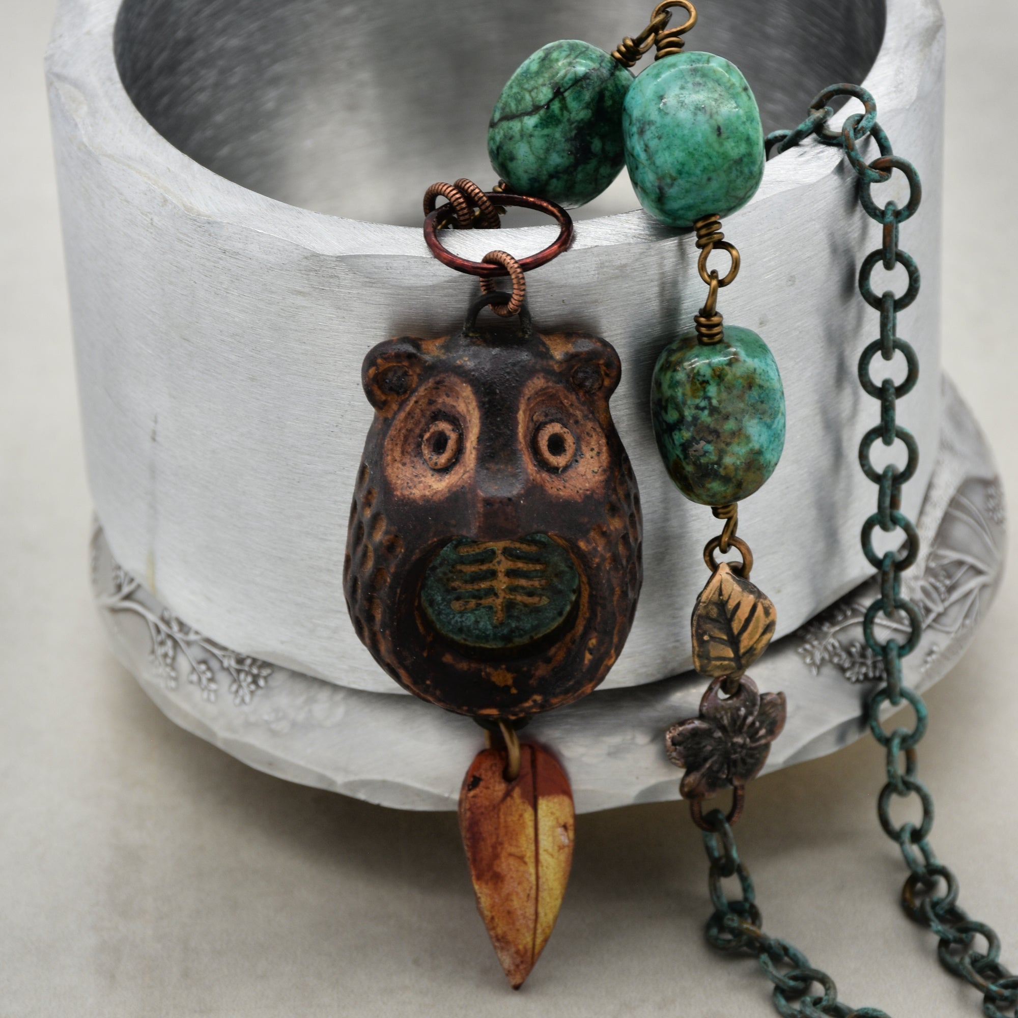 This image shows a bracelet. It features a chain with green beads and a pendant that resembles a bear's face with a small decorative leaf hanging from it. The design and length are consistent with what you would expect from a bracelet.