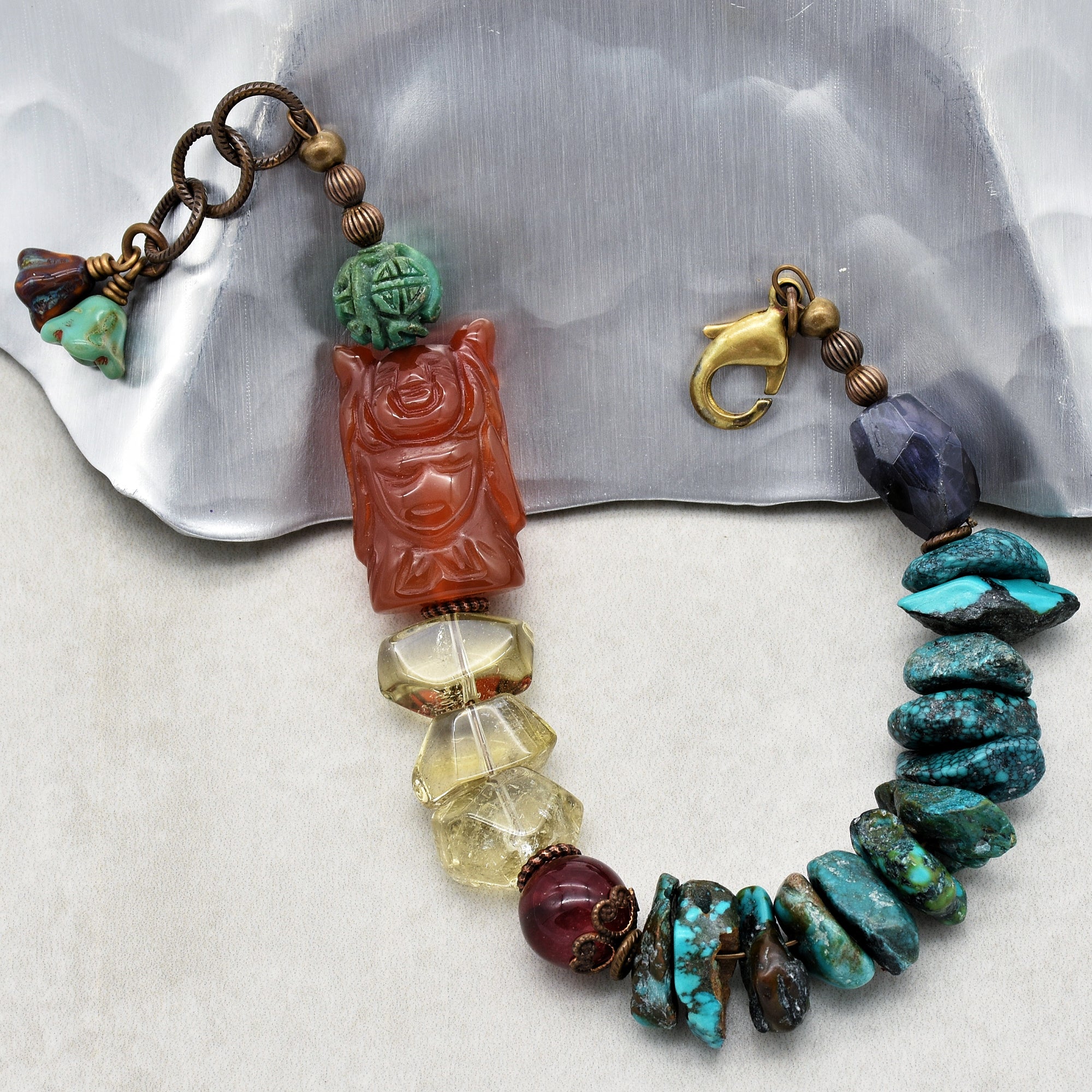 The image shows a bracelet. It features a variety of beads, including turquoise stones, a large carved red bead, a dark purple bead, and some translucent yellowish beads, all connected with a metal clasp.