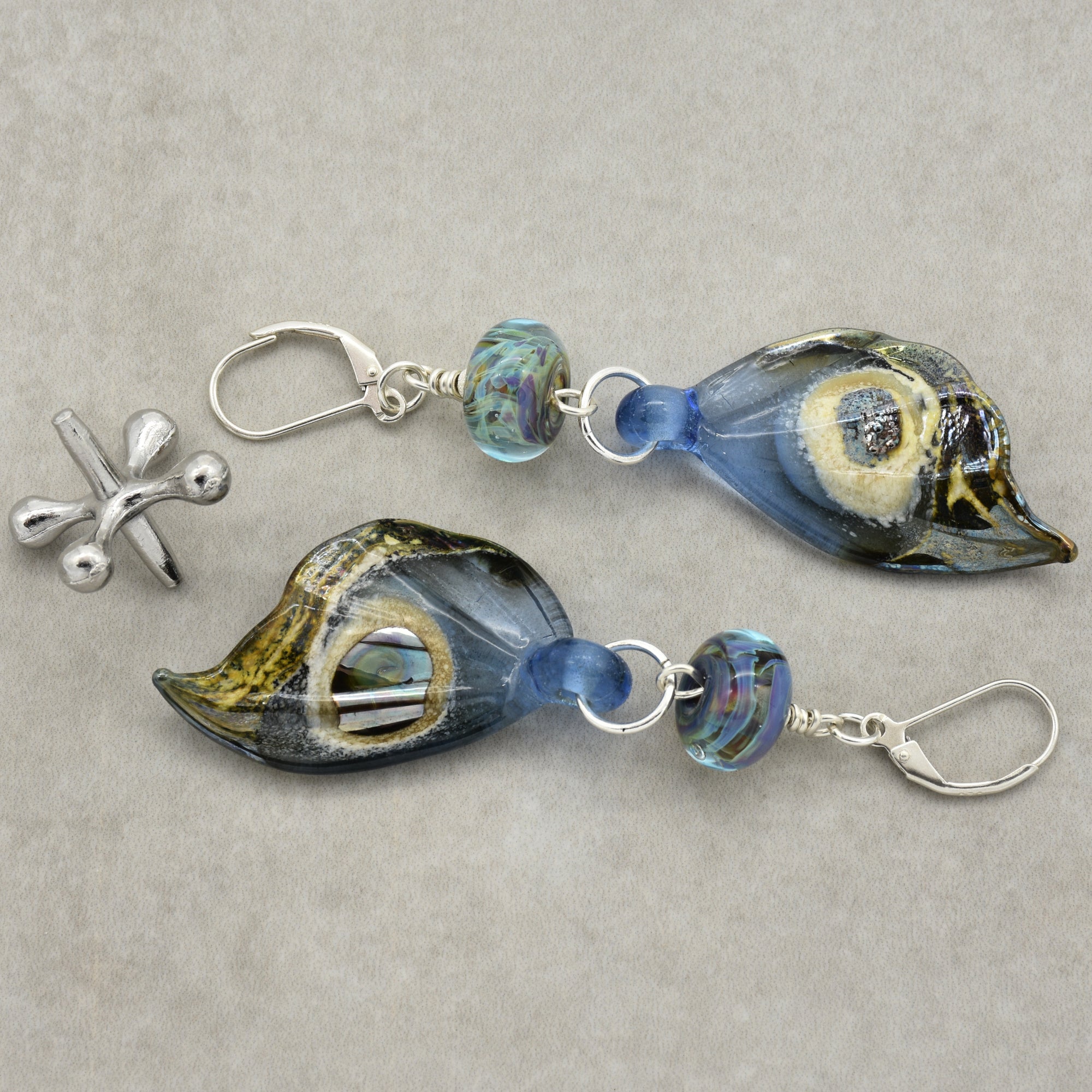 The image shows a pair of earrings. They are made with decorative glass pieces that resemble leaves or abstract shapes, combined with blue and multicolored beads and silver hooks.