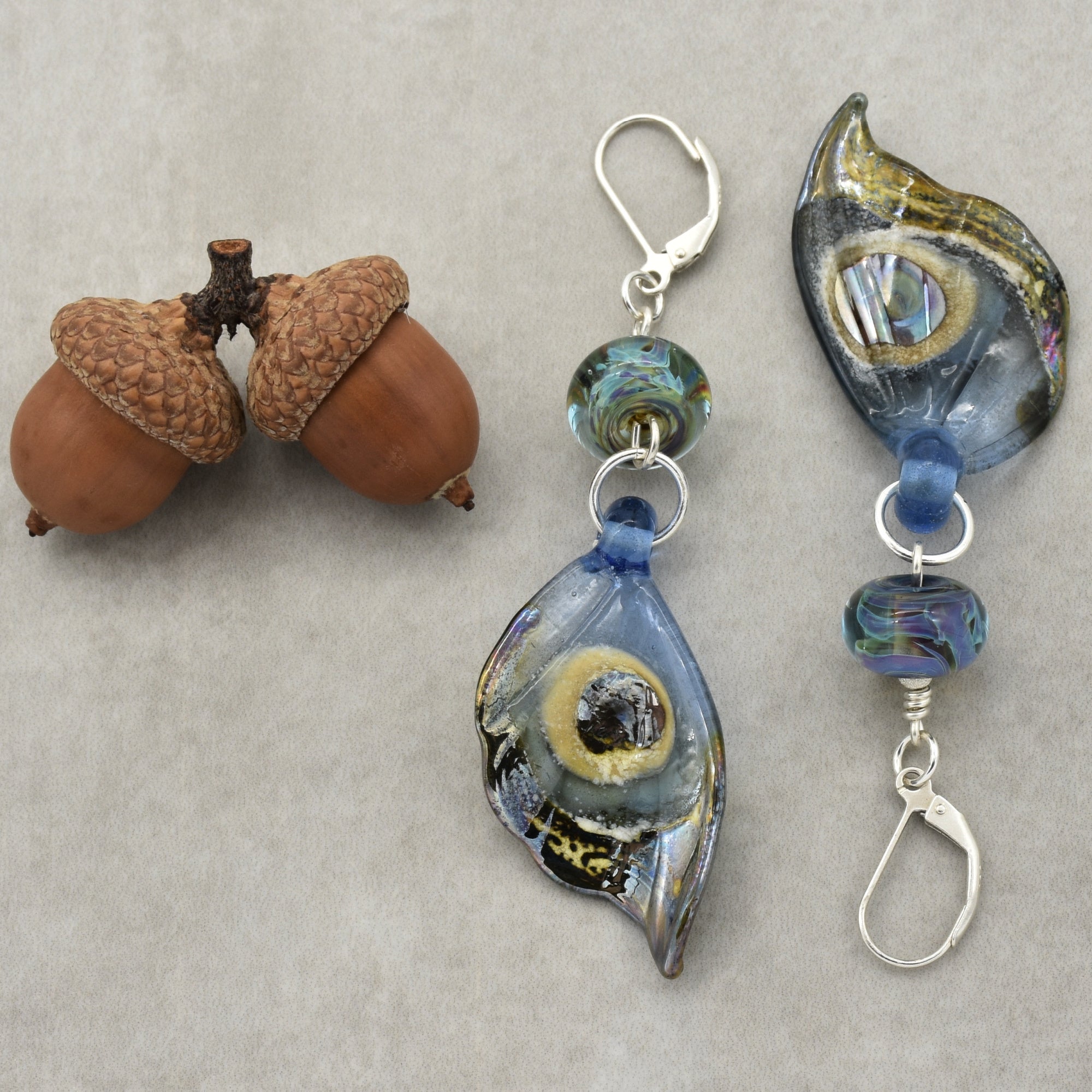 This image shows a pair of dangling earrings. They have a glass or resin pendant with blue and other colors, affixed to a clip-on or lever-back earring hook. The objects are clearly designed to be worn as earrings, not as a necklace or bracelet.