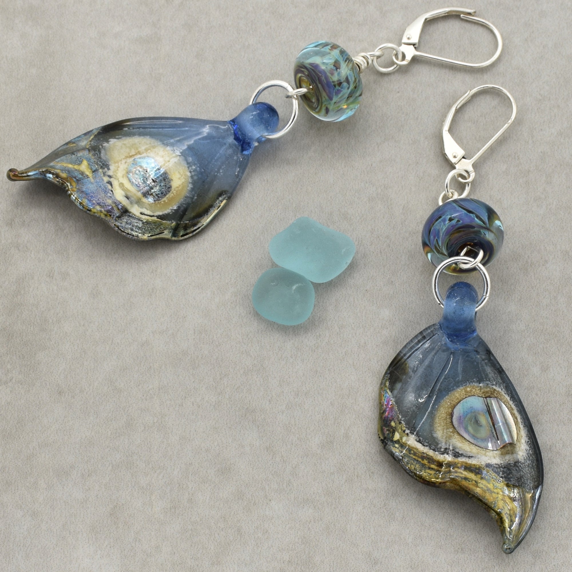 The image shows a pair of earrings. They have silver hooks and feature decorative glass or resin pieces shaped somewhat like leaves or petals, with blue, gold, and iridescent accents. The earrings also include round beads with a swirled blue and green pattern.