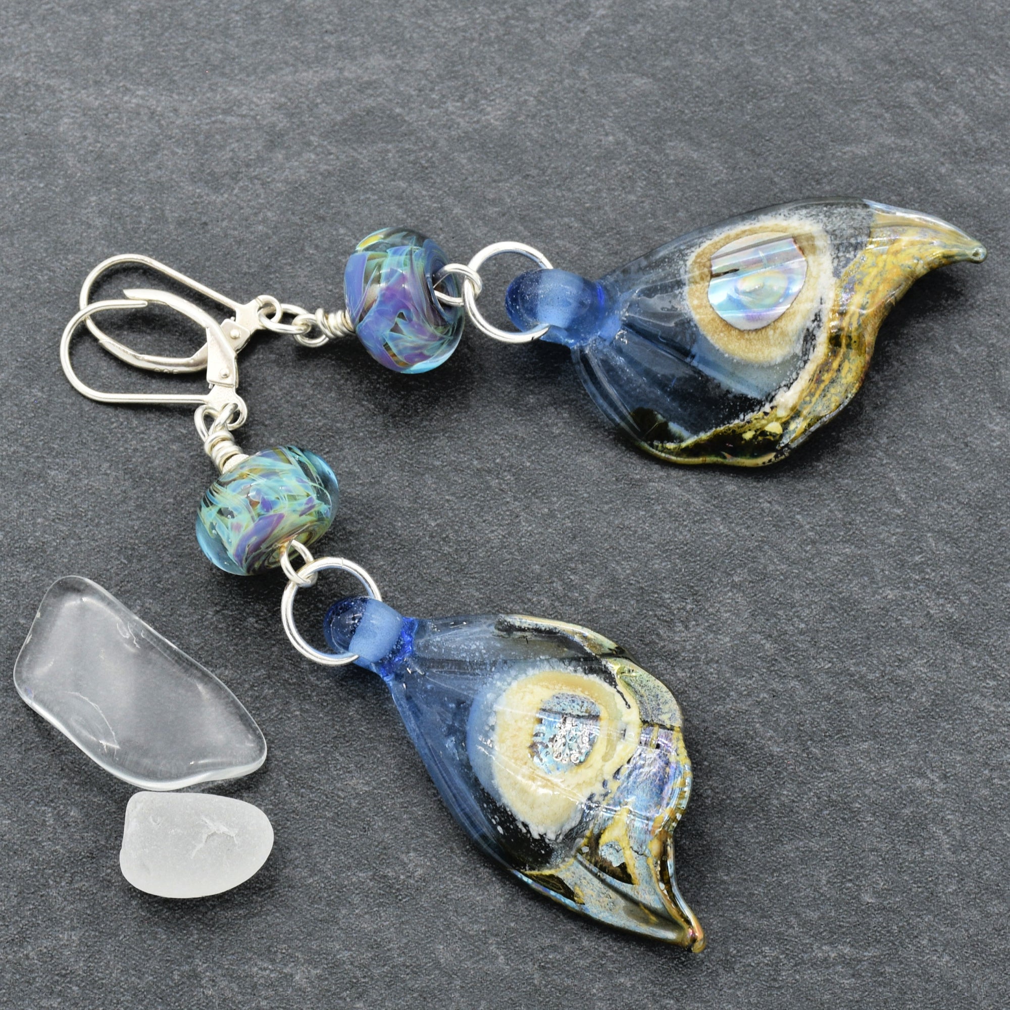 This image shows a pair of earrings. The jewelry consists of two matching pieces with hooks at the top for wearing in pierced ears. Each earring has decorative elements including colorful beads and glass or resin pendants shaped like stylized leaves or feathers.
