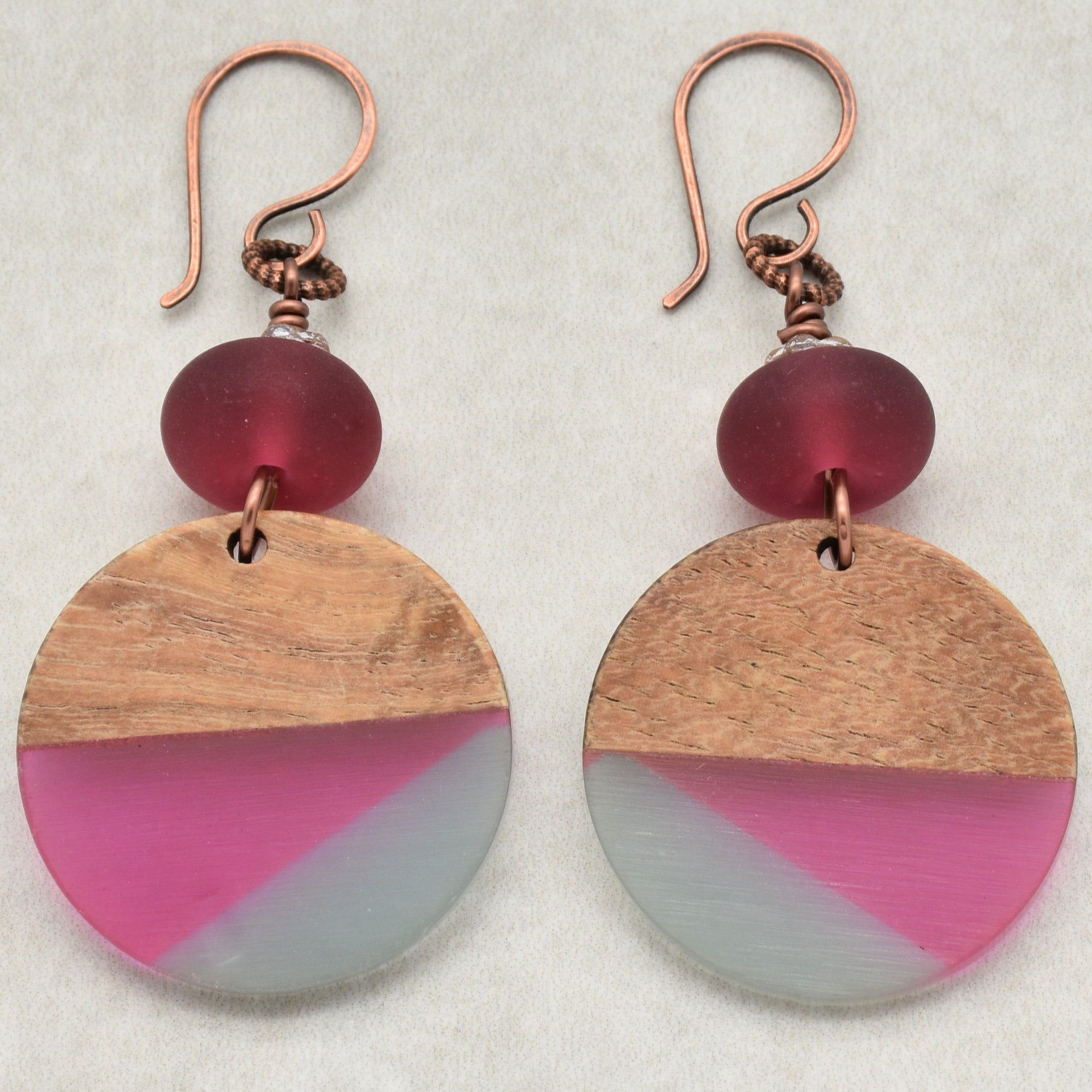 The image shows a pair of earrings. They have a circular wooden and colored design with a red bead above the circle and copper hooks for wearing.