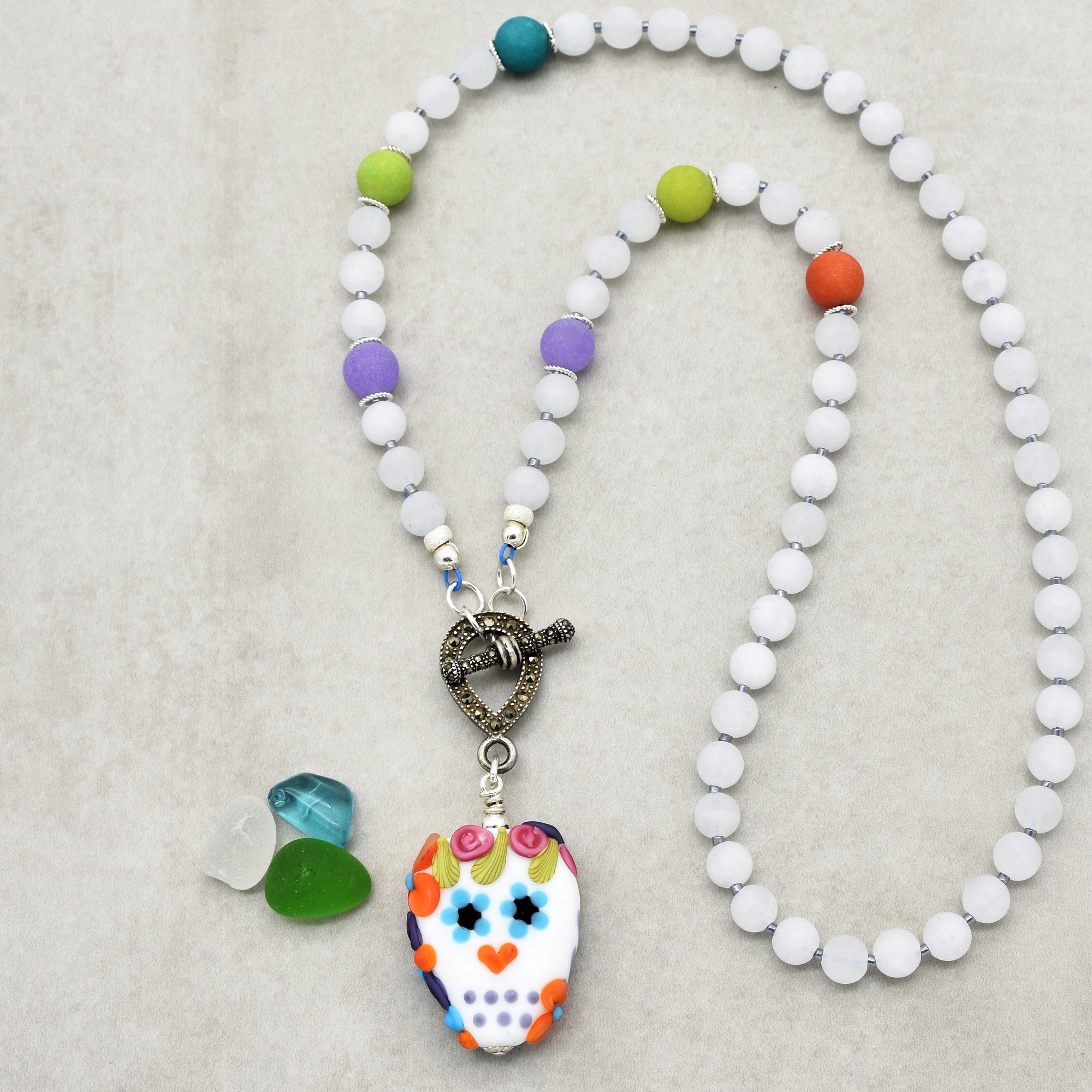 The image shows a necklace. It features a string of beads with a decorative clasp and a pendant shaped like a colorful skull. The beads are mostly white with some colored beads interspersed.