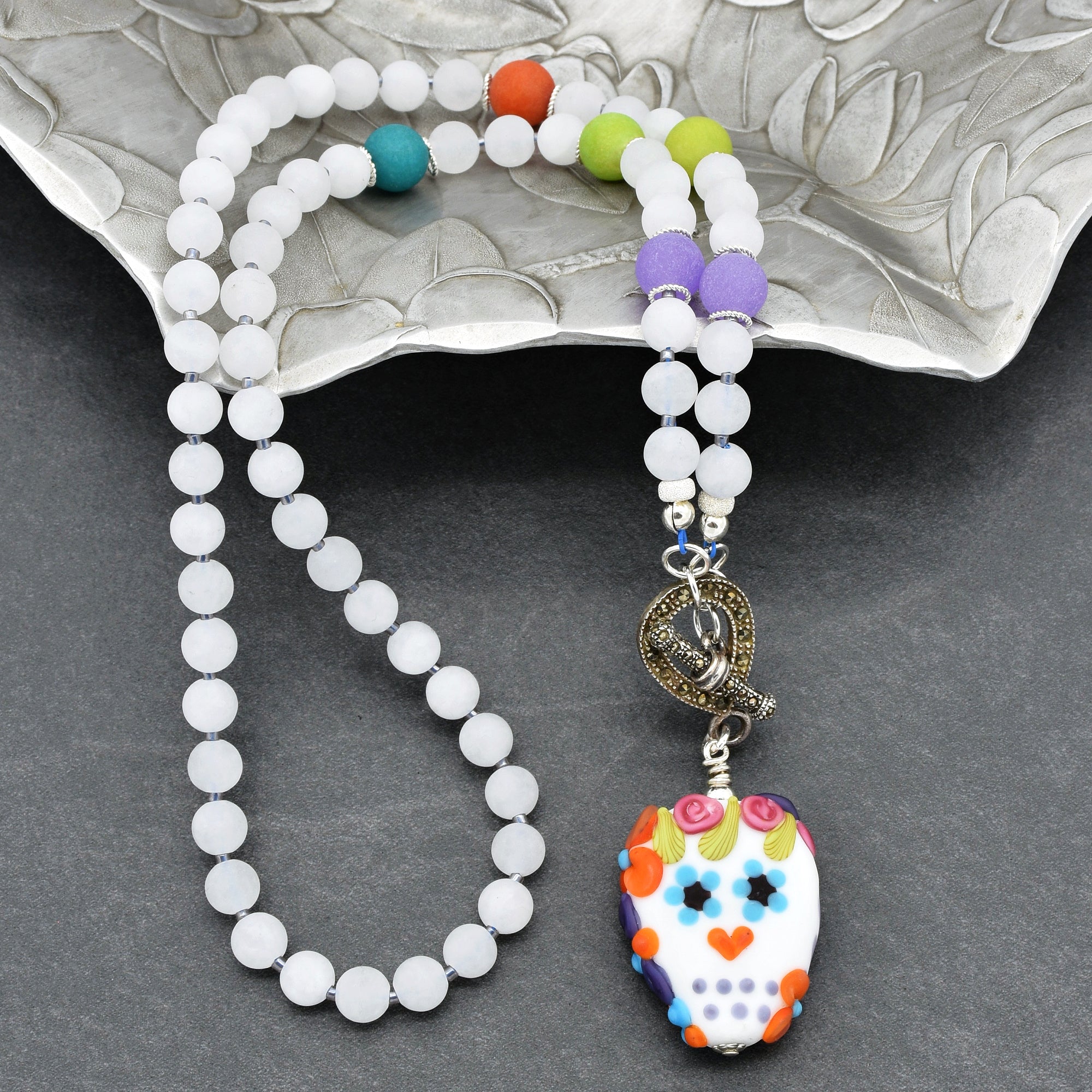 This image shows a necklace. It is made of white beads interspersed with a few larger colorful beads, and it features a decorative pendant with a colorful skull design. The presence of a clasp also suggests it is a necklace.