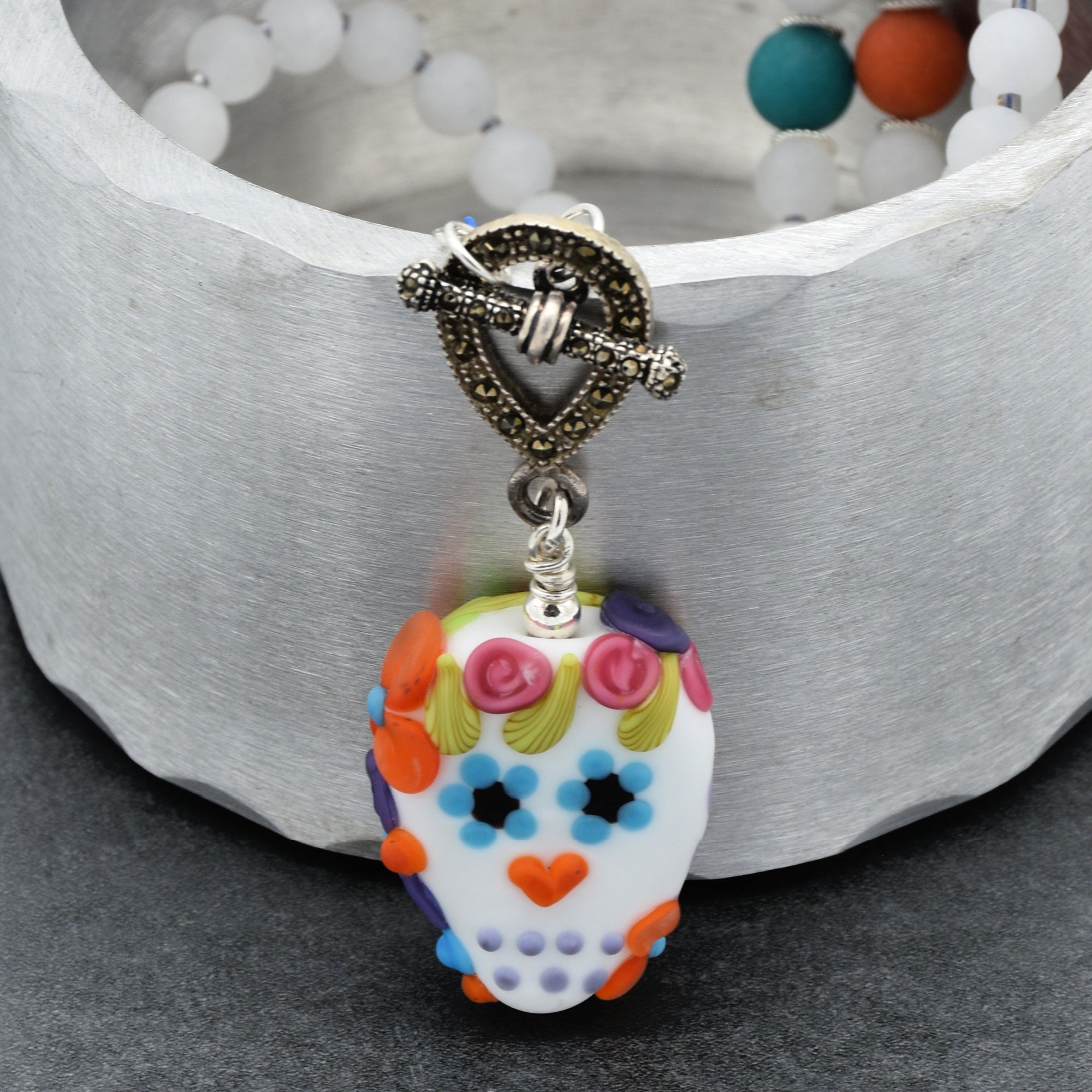 This image shows a necklace. The skull-shaped pendant with colorful floral designs is attached to a clasp that is part of a beaded necklace visible in the background.