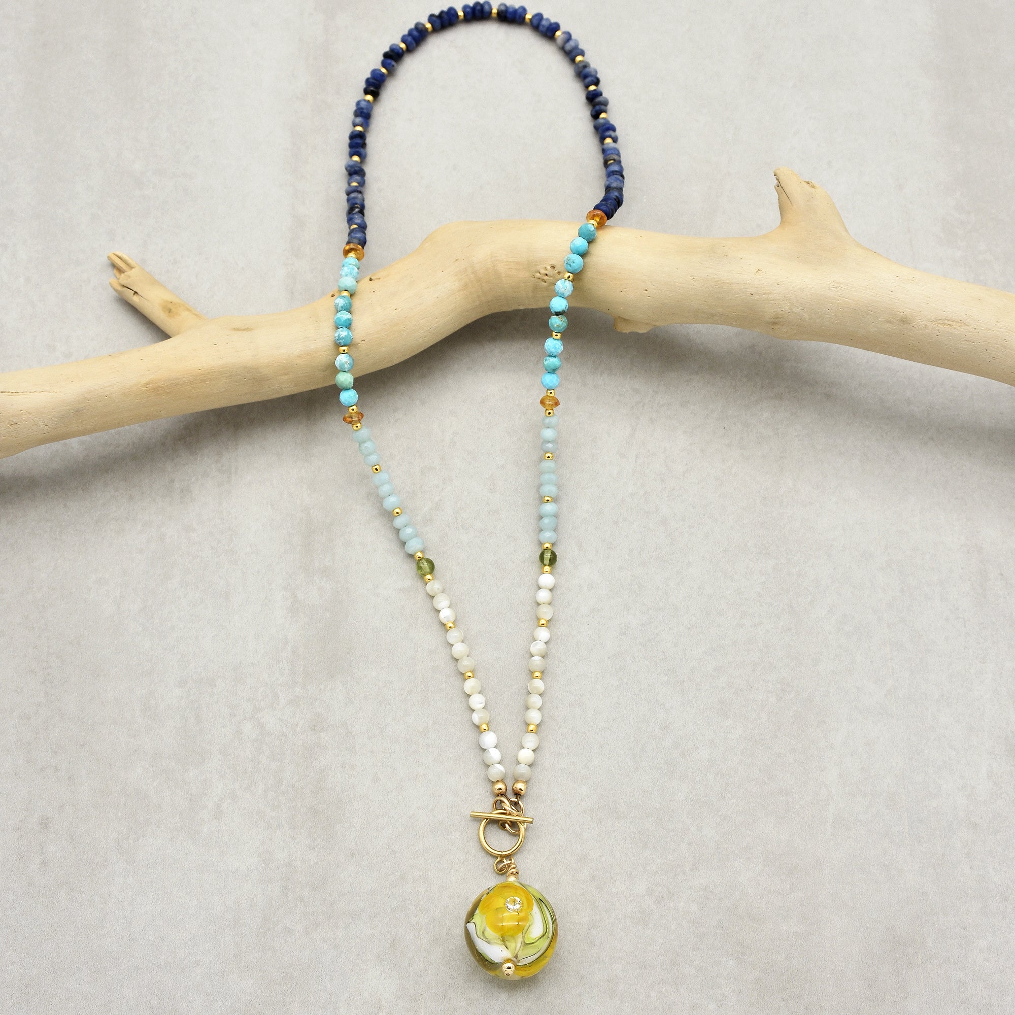 The image shows a necklace. It consists of beads in various colors including blue, turquoise, light blue, white, and small gold beads. It has a decorative pendant with a yellow and green design hanging at the center. The necklace appears to have a toggle clasp.