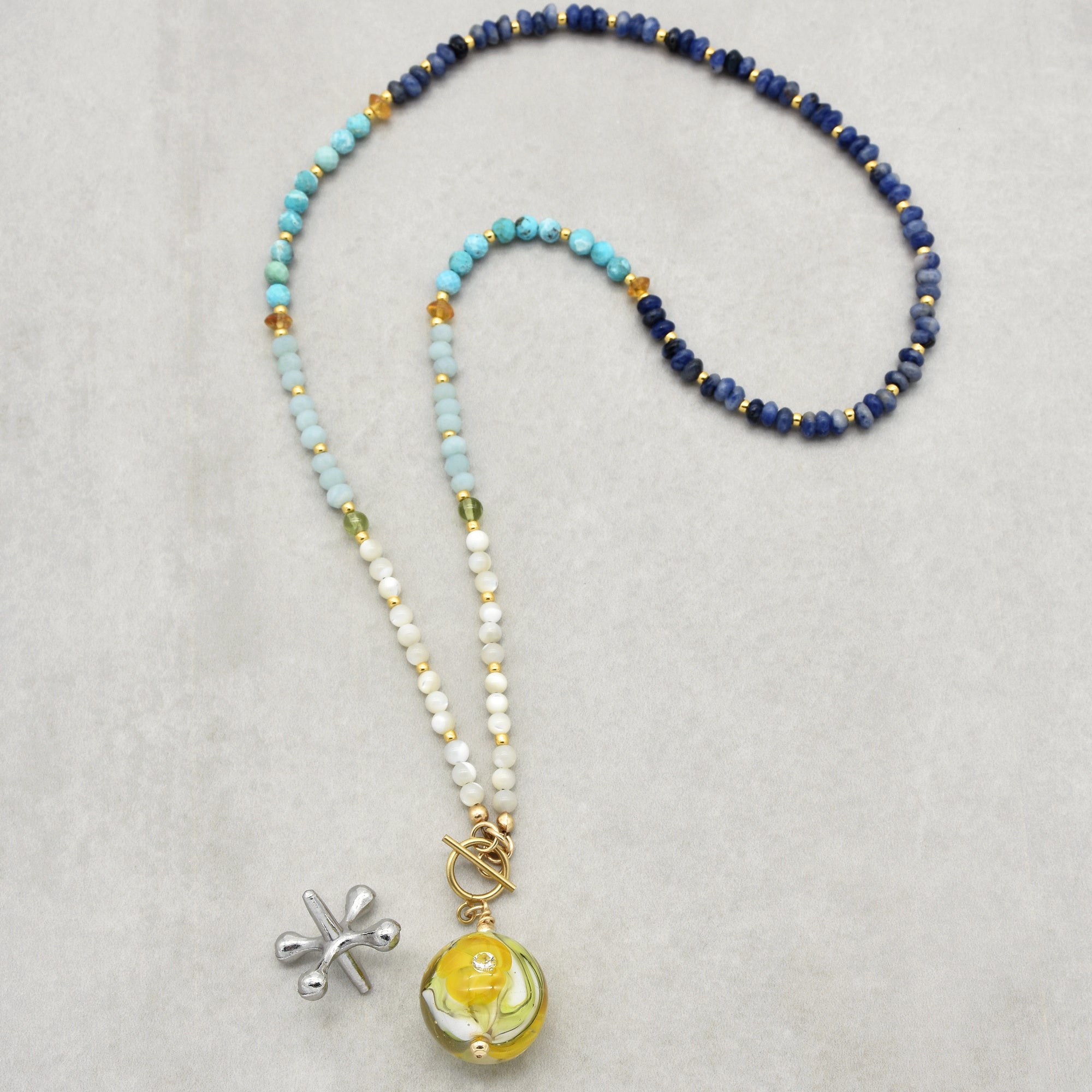This image shows a necklace. It has a beaded design with various colors, including blue, turquoise, white, and green beads, accented with small gold beads. The necklace features a round glass pendant with a yellow flower design and a round clasp nearby.