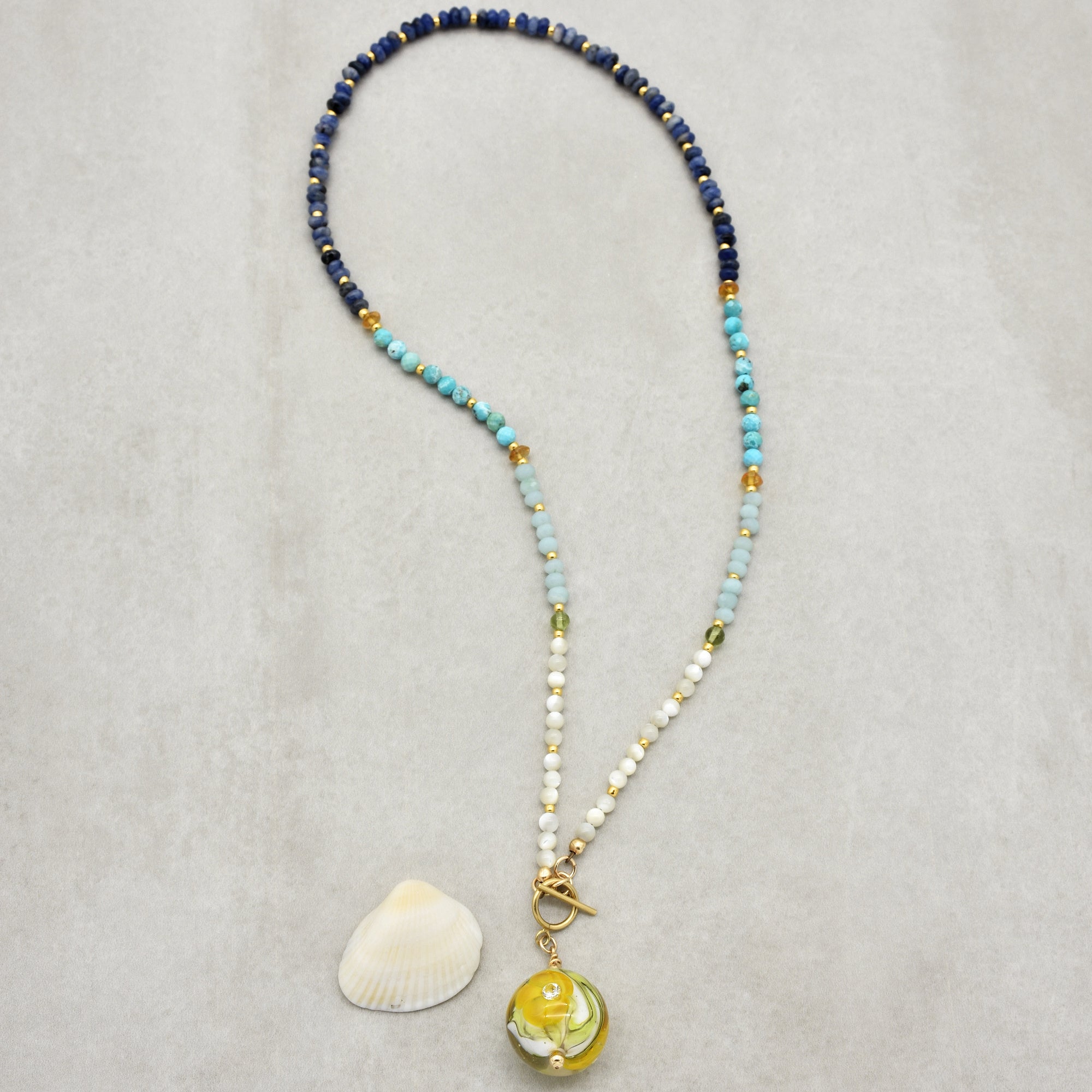 This image shows a necklace. The jewelry features multicolored beads in shades of blue, white, and gold, with a yellow and green round pendant at the end. Additionally, the necklace is laid out next to a small seashell.