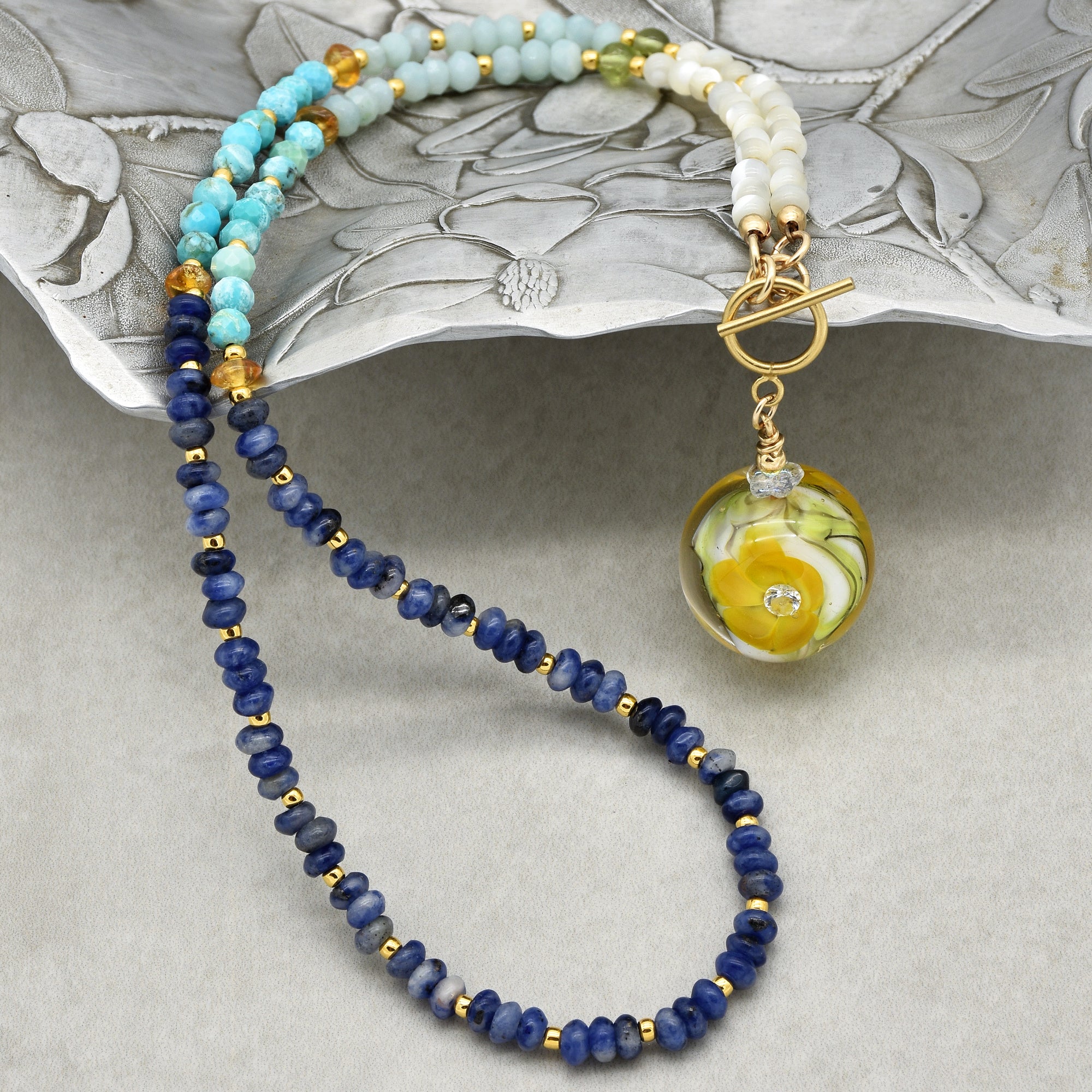 This image shows a necklace. The jewelry piece is made of various beads in shades of blue, white, and gold with a round pendant featuring a yellow floral design. The clasp also suggests it is meant to be worn around the neck.