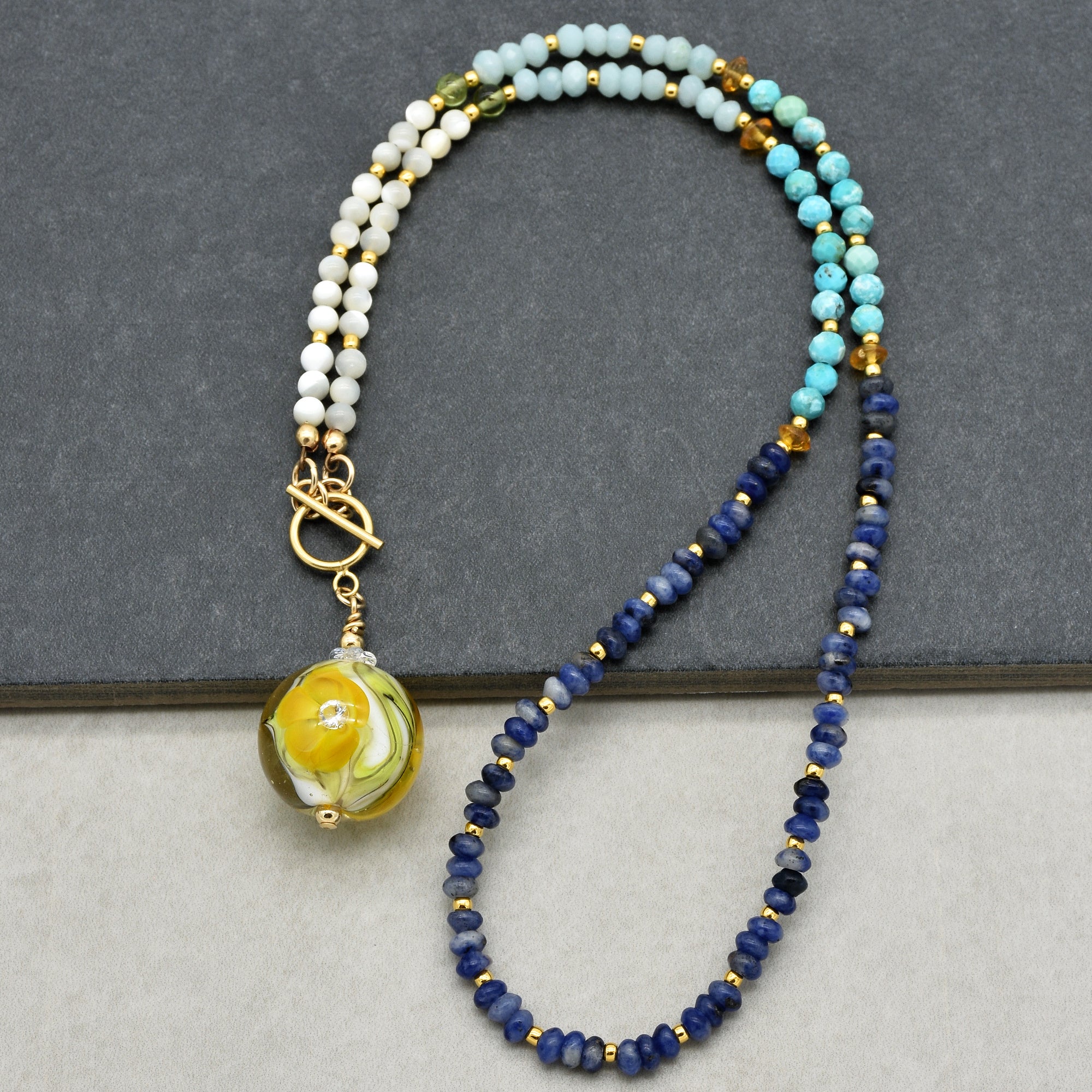 The image shows a necklace. The jewelry is composed of various beads in shades of blue, white, and turquoise with gold spacers, and it features a round yellow pendant. The necklace has a toggle clasp visible on the left side of the image.