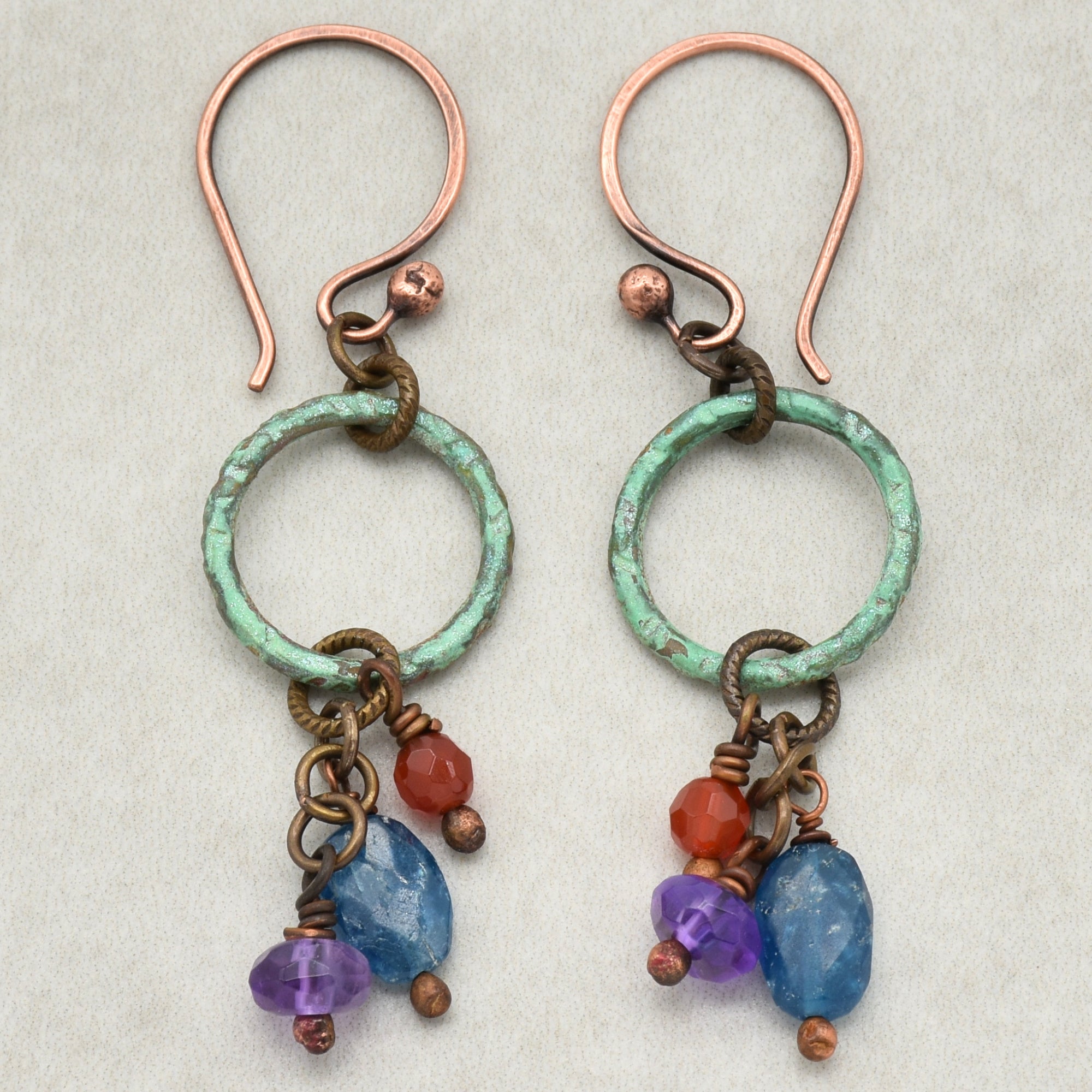 This image shows a pair of earrings. They have hooks for the ears at the top, with a green circular ring and dangling colored beads (red, blue, and purple) below.