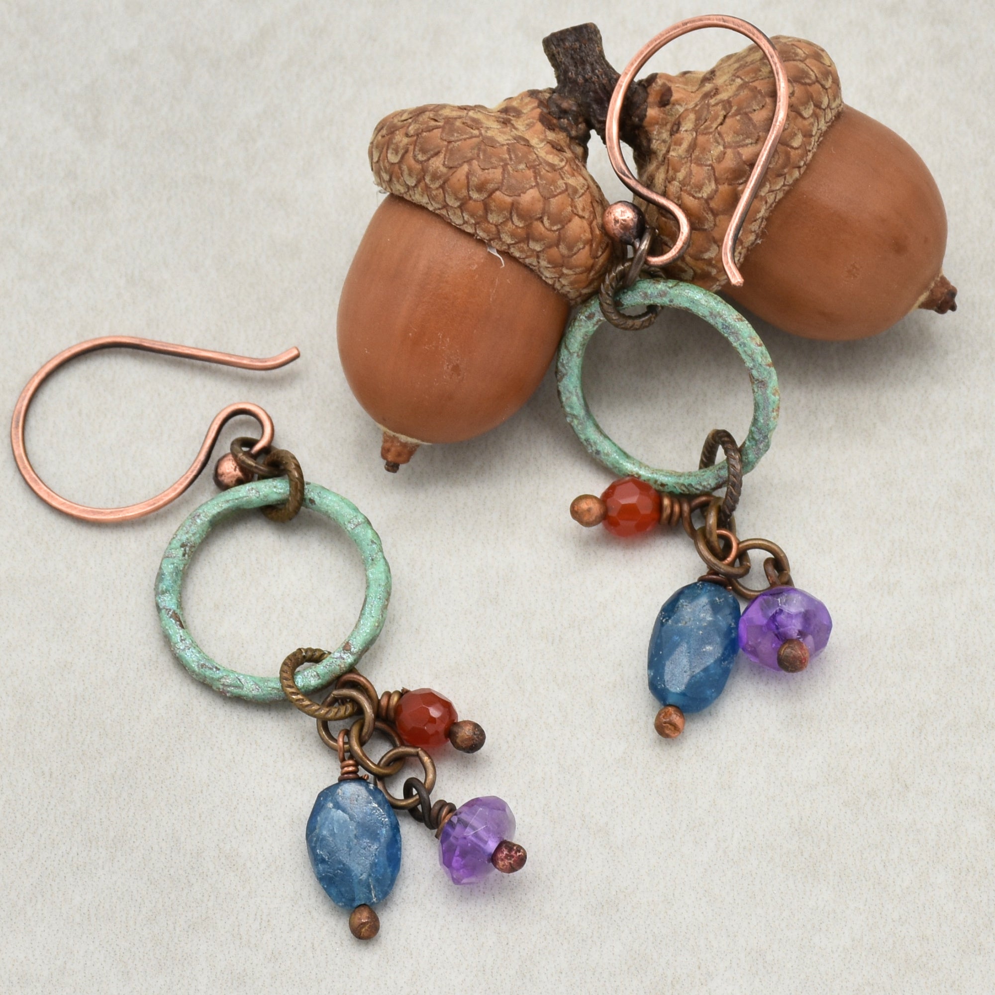 The image shows a pair of earrings. They have copper hooks and feature a circular greenish-blue ring with small hanging beads in blue, purple, and red colors.