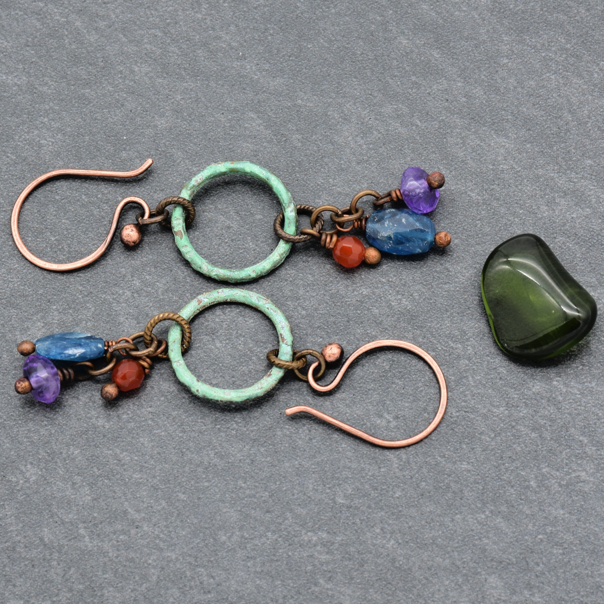 The image shows a pair of earrings. They have copper hooks with circular green rings and several small colorful beads attached.