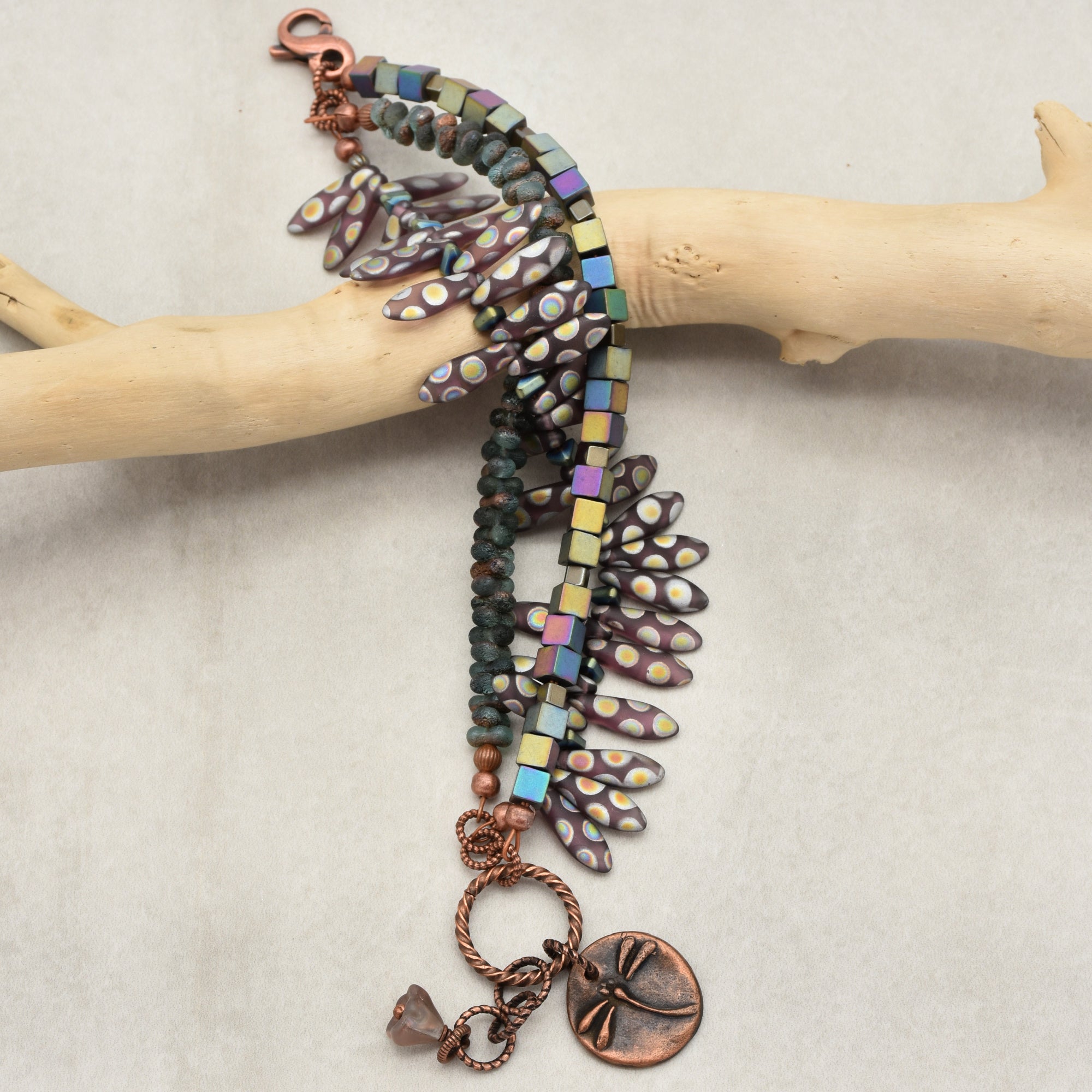 This image shows a bracelet. It features a clasp, decorative beads including iridescent cube-shaped beads, elongated beads with a dotted pattern, and a copper charm with a dragonfly design. The bracelet is draped over a piece of wood for display.