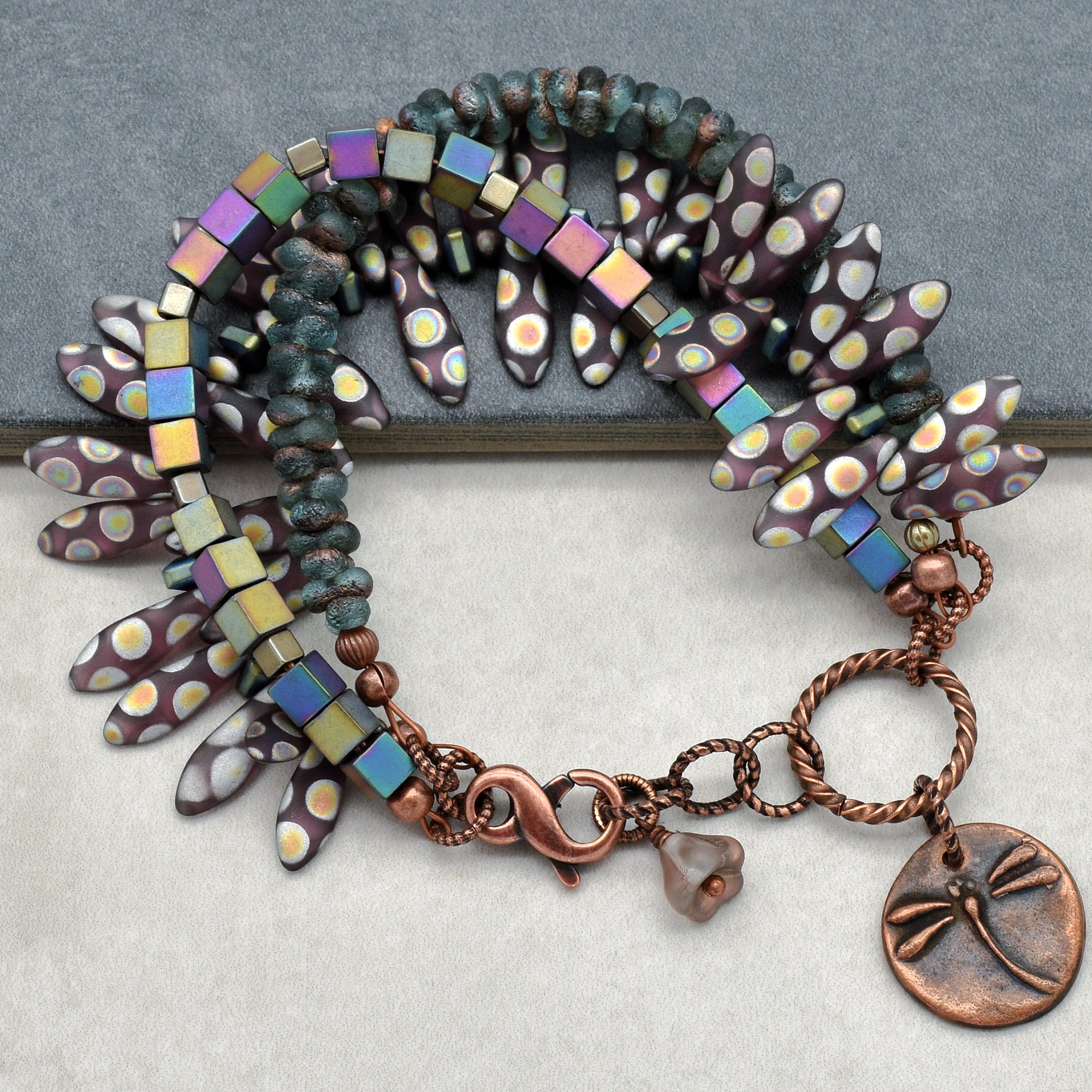 This image shows a bracelet. The jewelry piece is composed of textured beads, square iridescent beads, and petal-like elements with a dotted pattern, arranged in a circular form typical of bracelets. Additionally, it has a clasp and a charm with a dragonfly design, which are common features of bracelets.