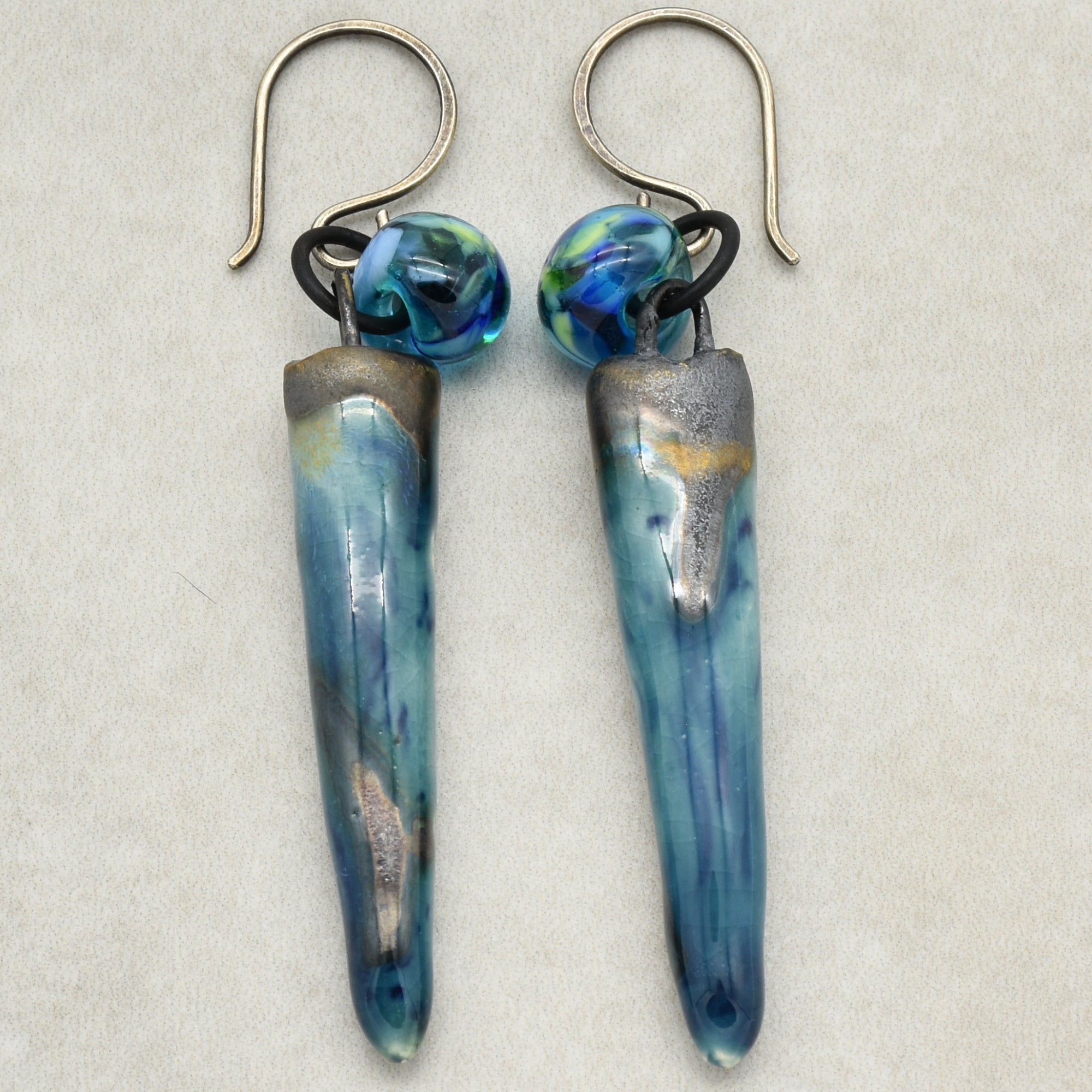 The image shows a pair of earrings. They have hook-style ear wires and blue elongated decorative elements hanging from them with a small round bead at the top.