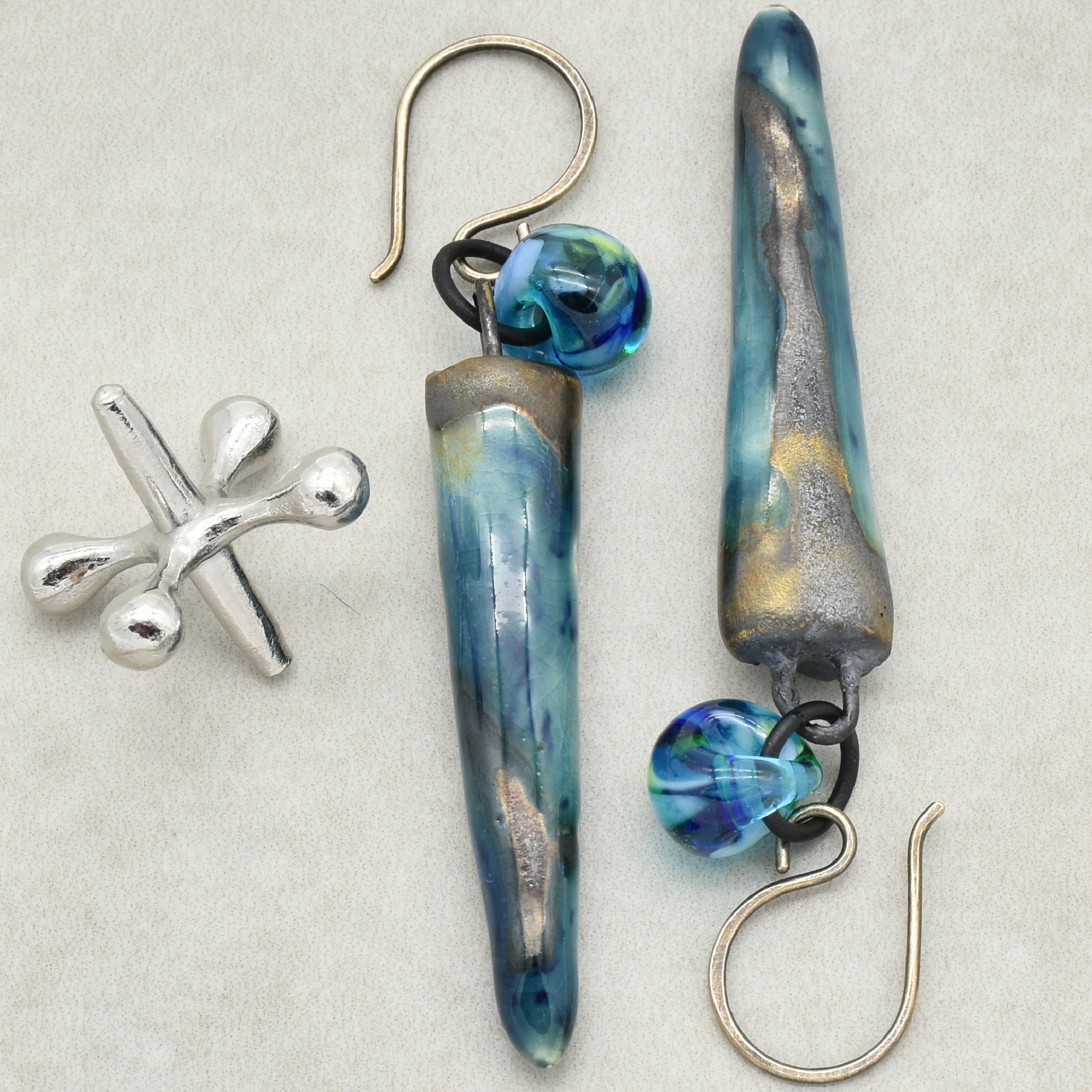 The image shows a pair of earrings. They have a long, tapered blue and metallic design with a small round blue bead near the hook part that goes through the ear. There is also an additional small metallic piece with a unique shape on the left side of the image, but the main items appear to be earrings.