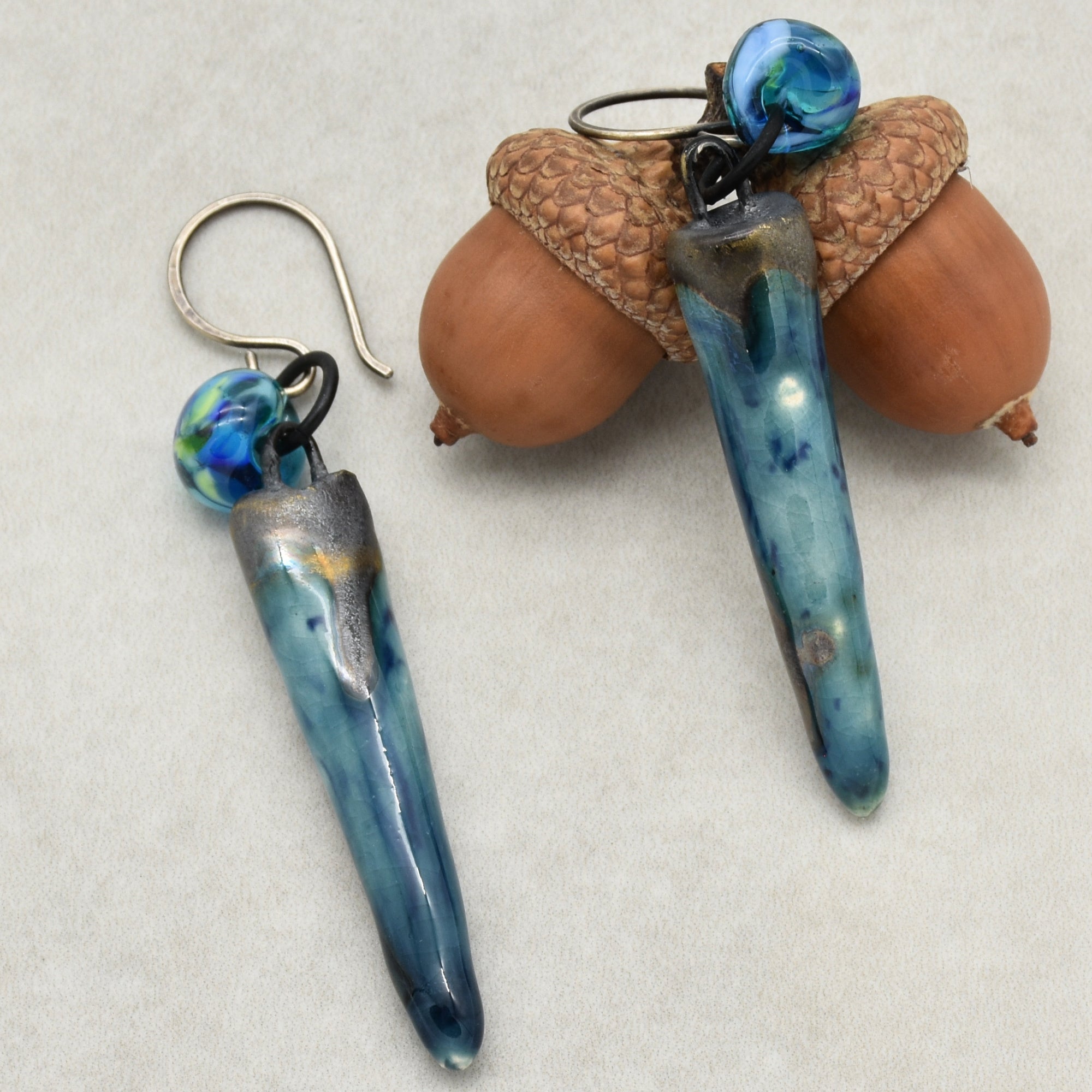 The image shows a pair of earrings. They have hooks for pierced ears and feature elongated blue beads with smaller decorative beads above them. One of the earrings is displayed on top of two acorns.