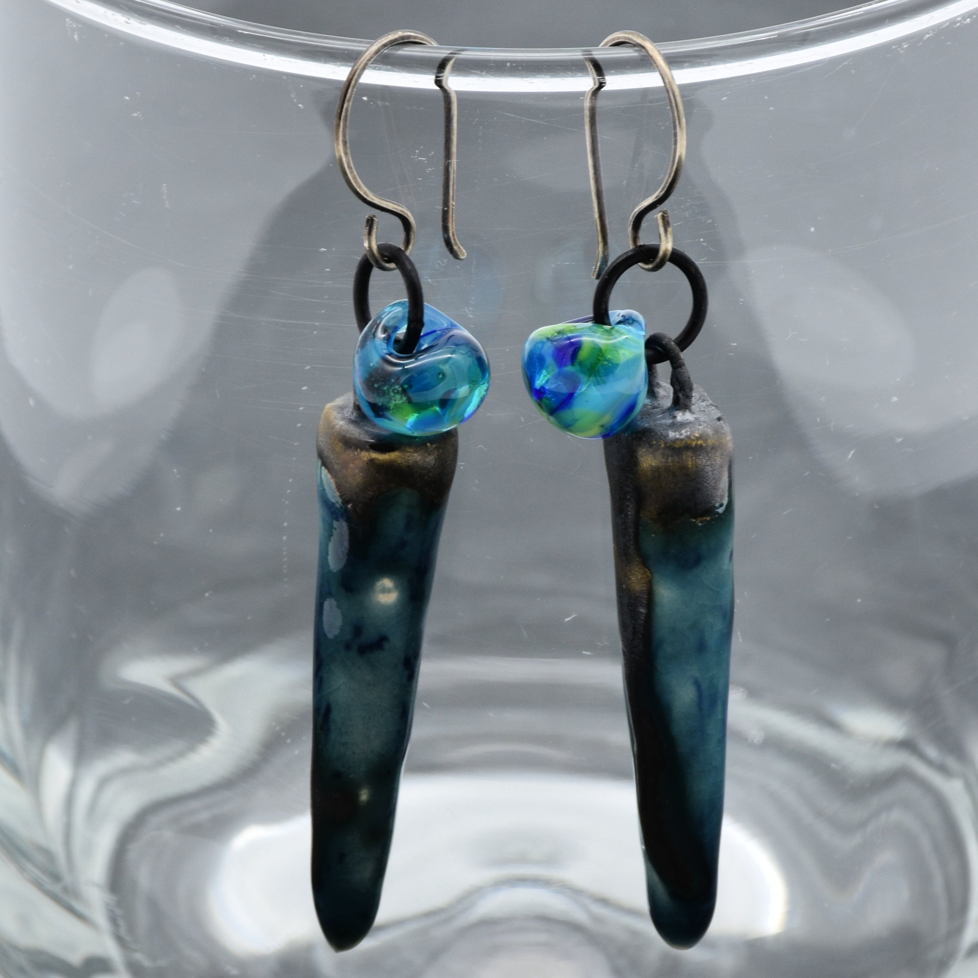 The image shows a pair of earrings. They have hooks for piercing and feature elongated, irregular shapes with blue and green tones as part of the design.
