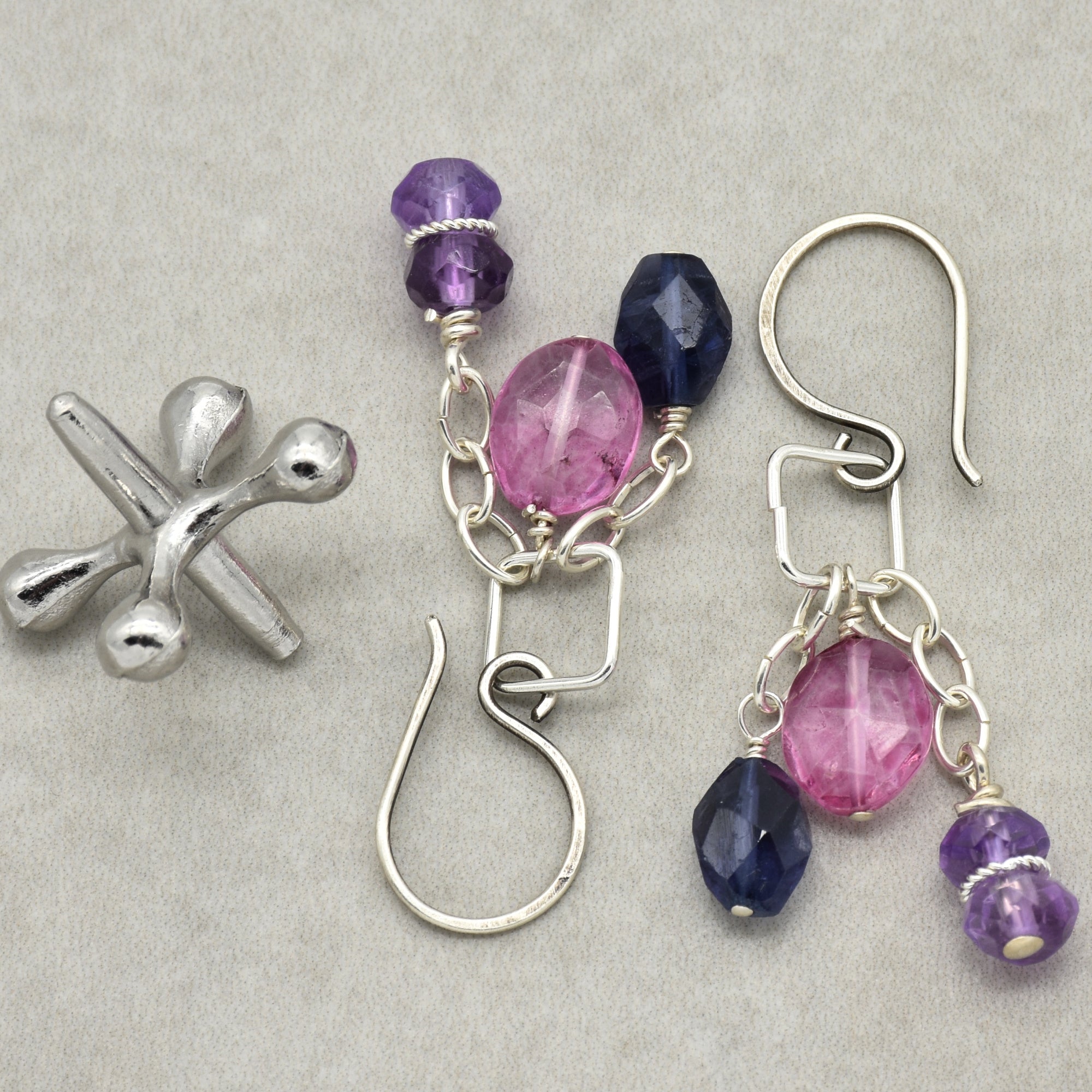 The image shows a pair of earrings. They have hooks for piercing and are decorated with purple, pink, and dark blue beads hanging from small silver chains. There is also a silver metallic object shaped like a jack next to the earrings, but it is not part of the jewelry type in question.