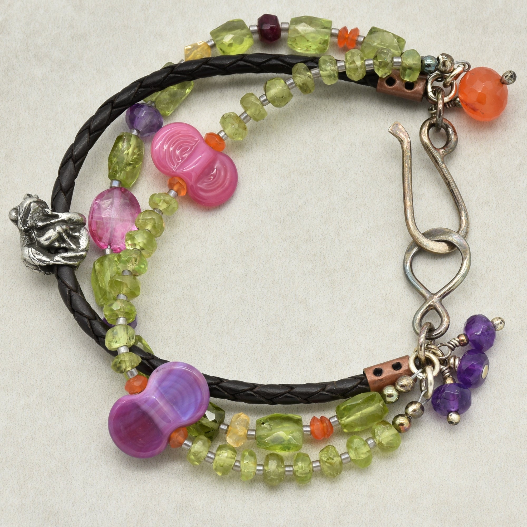 This is an image of a bracelet. It features multiple strands with various beads including green, purple, and pink ones, along with a braided leather strand. The closure is a metal hook clasp. The size and layout of the jewelry piece indicate that it is a bracelet rather than a necklace or earring.