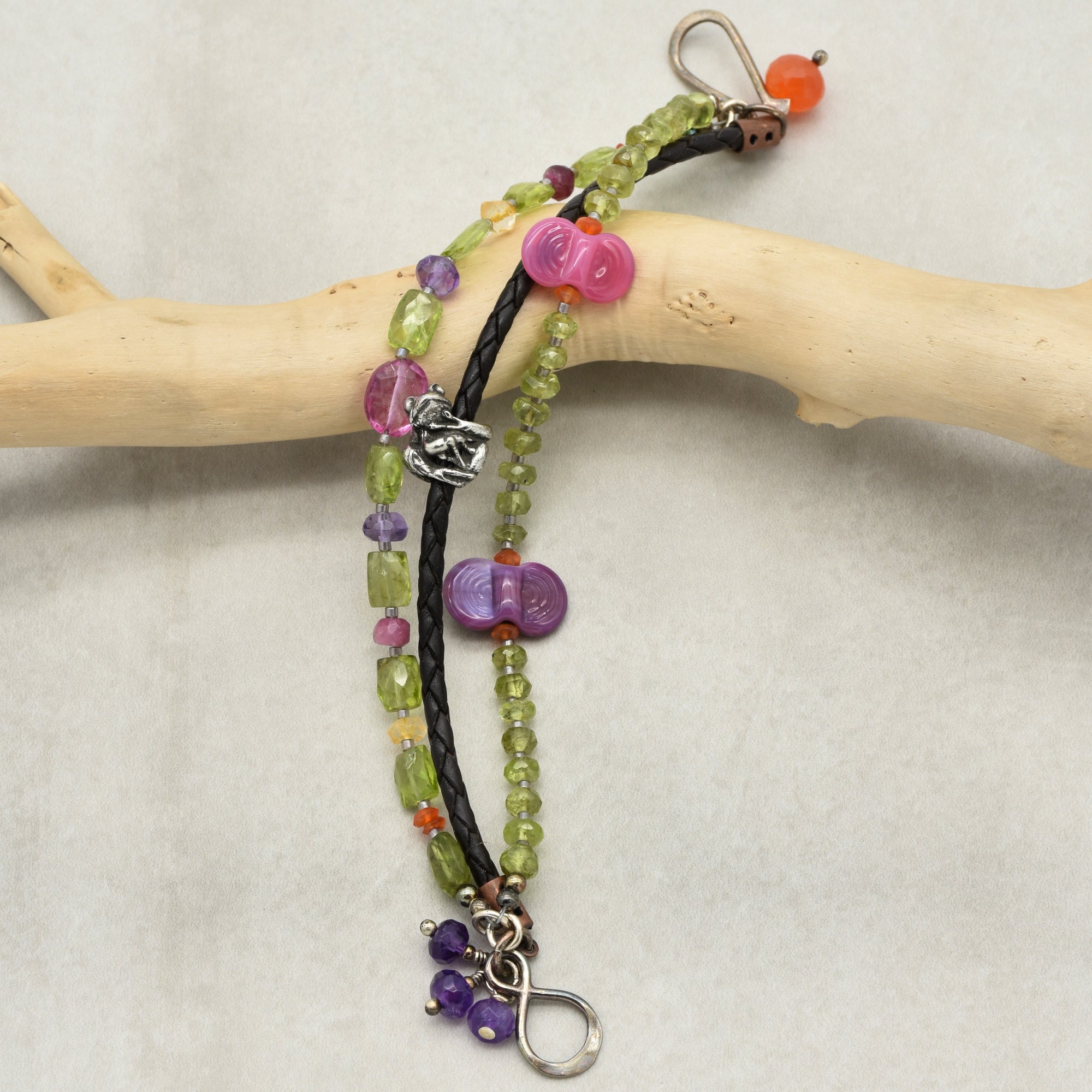 The image shows a bracelet. It features multiple strands with beads in various colors including green, purple, and pink, along with a braided black strand. The bracelet also has a clasp for fastening.