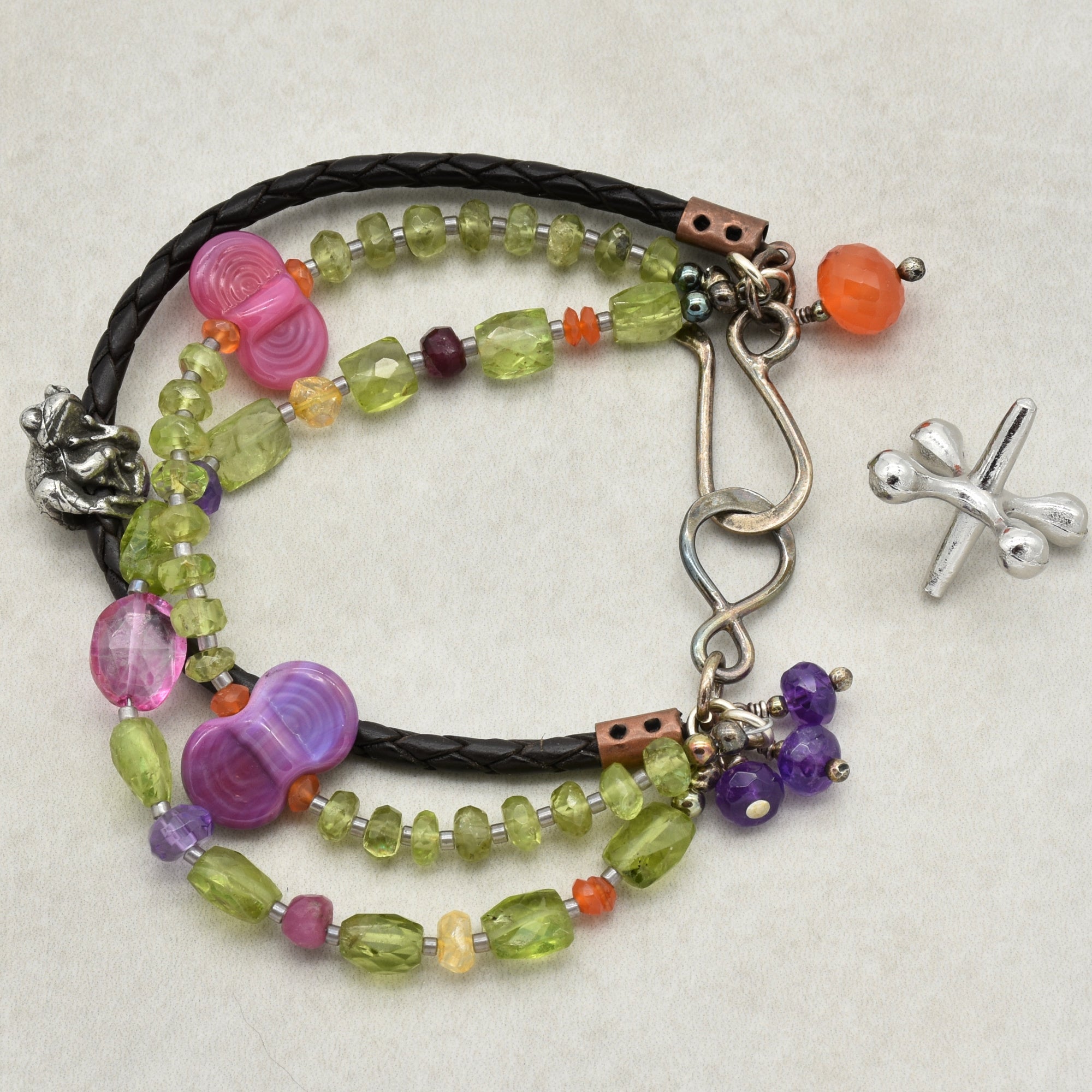 The image shows a bracelet. It features multiple strands with colorful beads, including green, purple, and pink tones. The bracelet also has a leather or faux leather braided strand and a metal clasp with small charms attached.