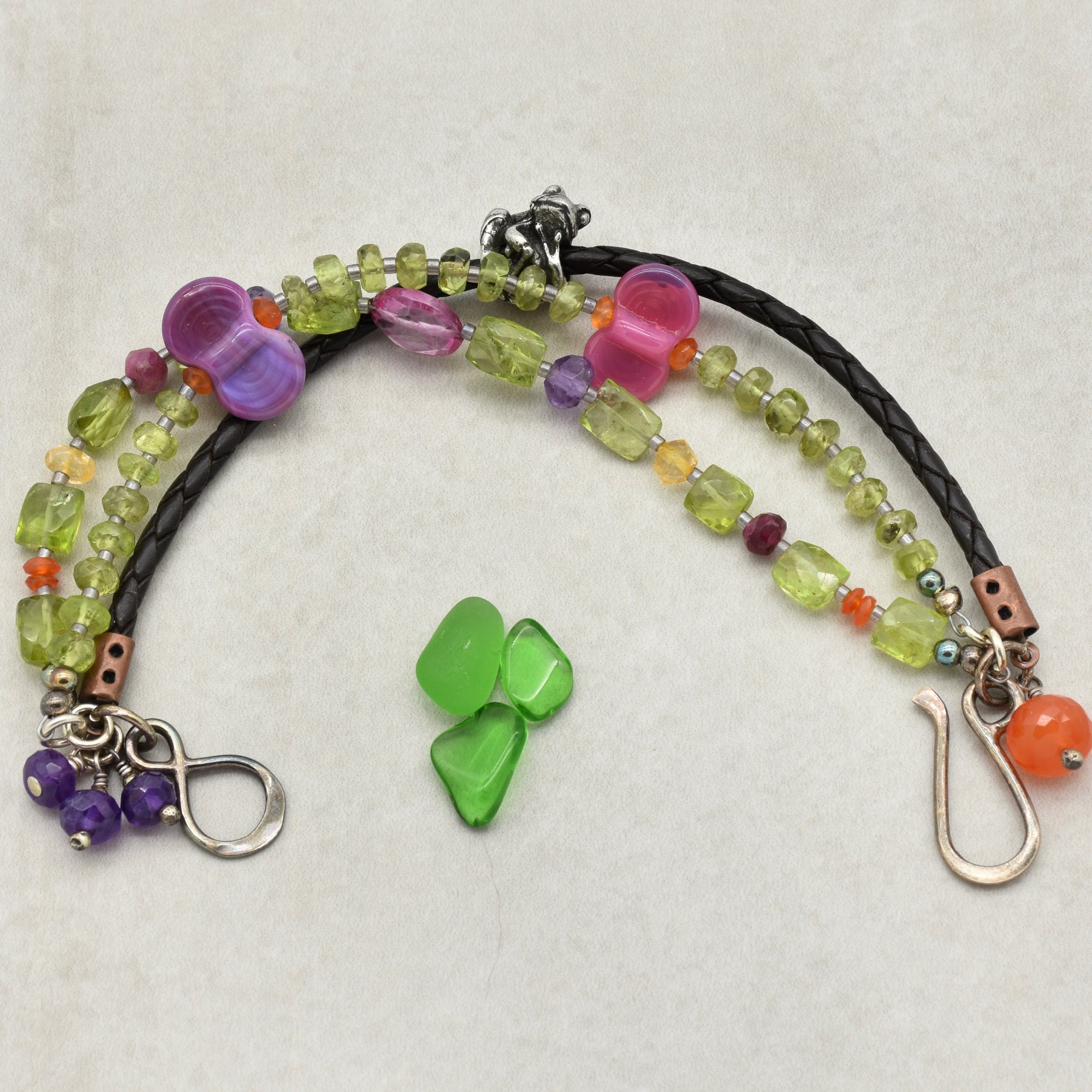 This image shows a bracelet. The jewelry piece has two strands, one with colorful beads including purple, pink, green, and orange, and the other appears to be a braided black cord. The ends have clasps that are typical of a bracelet closure.