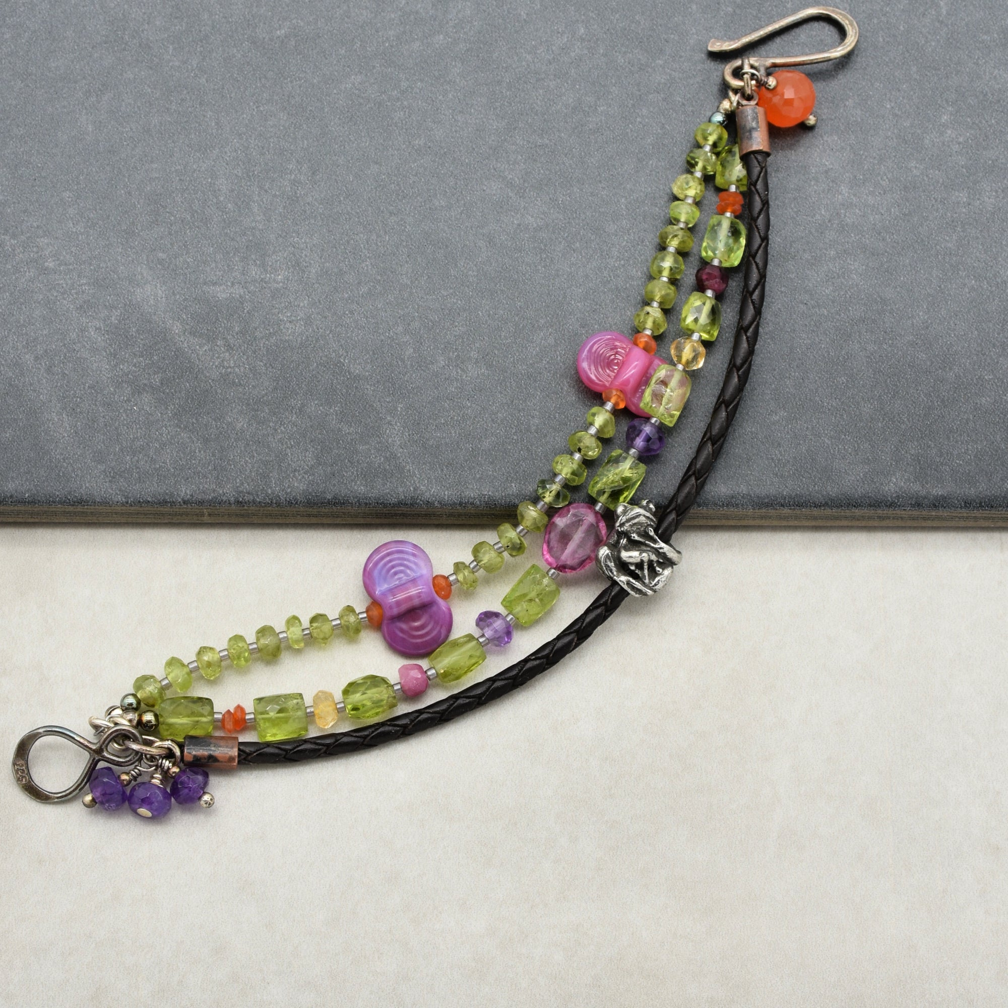 This image shows a bracelet. The bracelet features multiple strands with a combination of green, purple, pink, and orange beads, along with a braided black leather strap and a metal clasp.