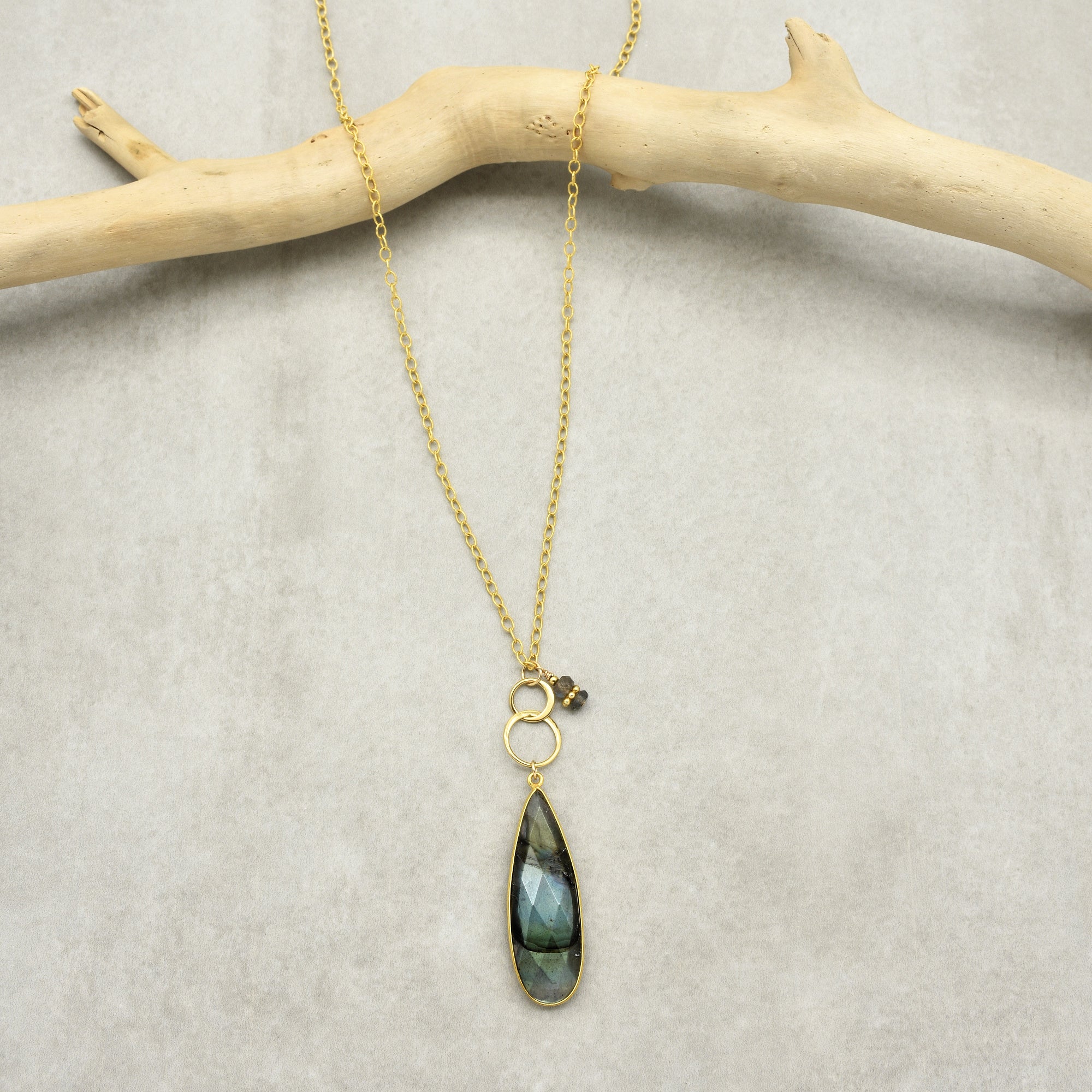 This image shows a necklace. It features a gold chain with a pendant that includes two small gold rings and a long, faceted dark gemstone, as well as a small cluster of dark beads or stones next to the rings.