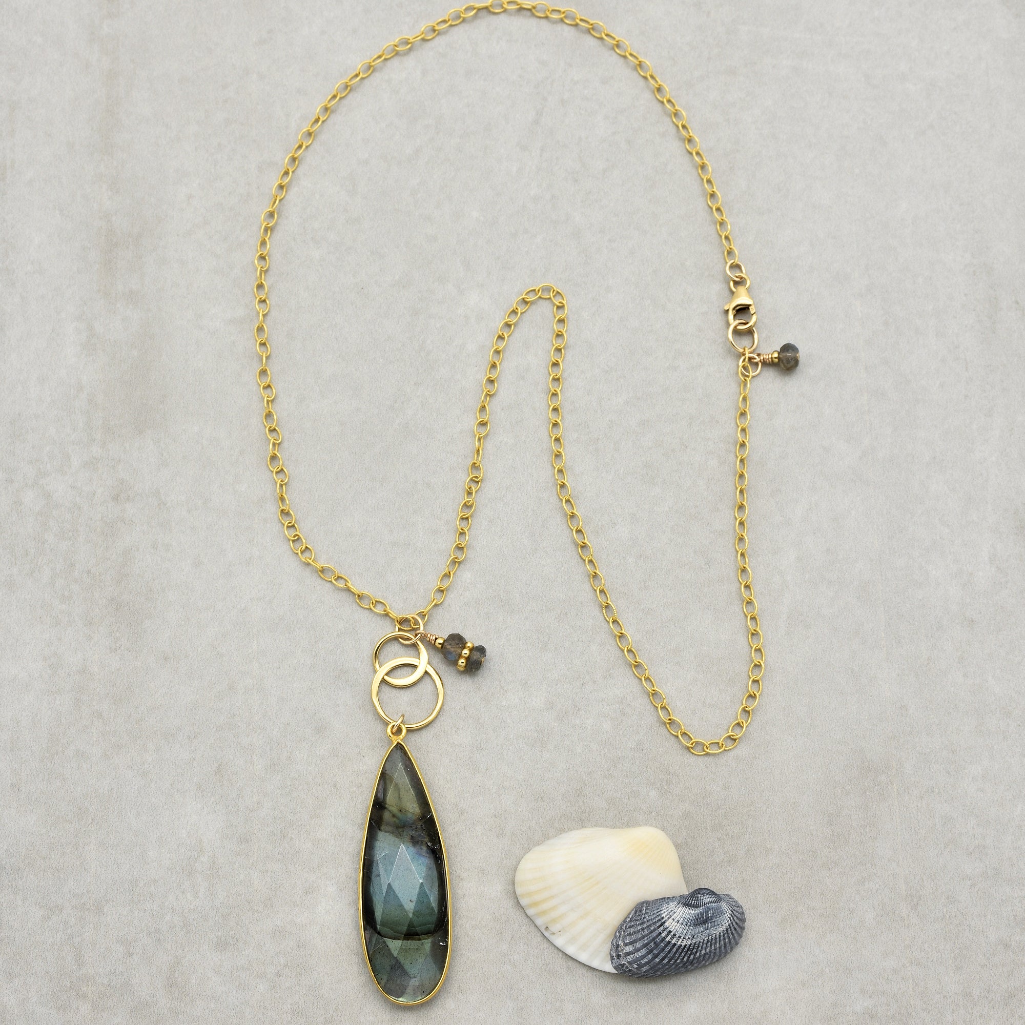 The image shows a necklace. It features a gold chain with a large teardrop-shaped pendant in shades of green and blue, along with two interlocking gold rings and small gemstone charms near the clasp and the pendant. There are also two seashells placed nearby for decoration.